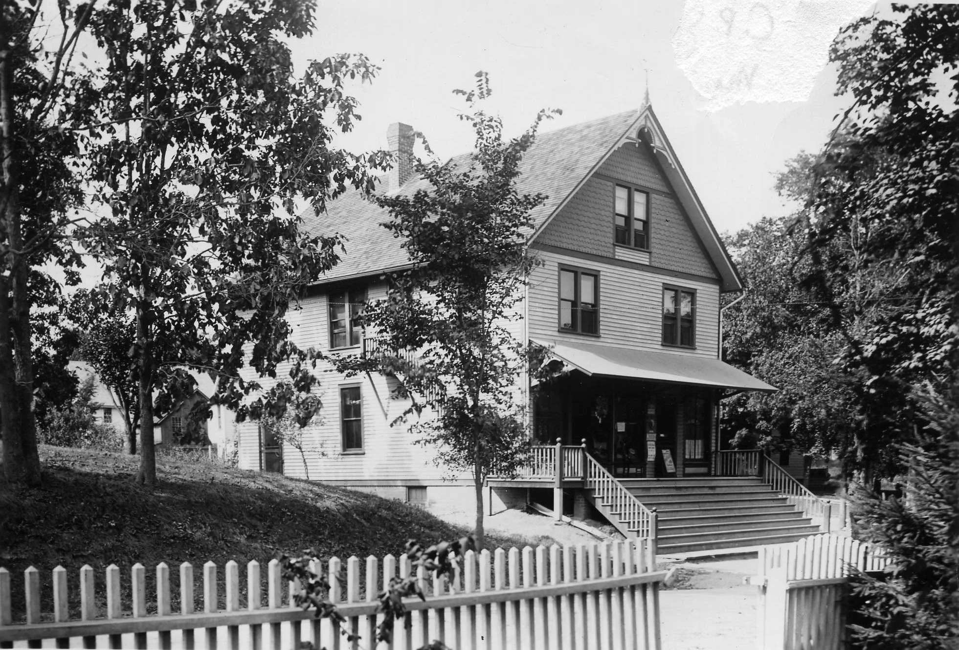 Historic house with a porch and trees, surrounded by a white picket fence.