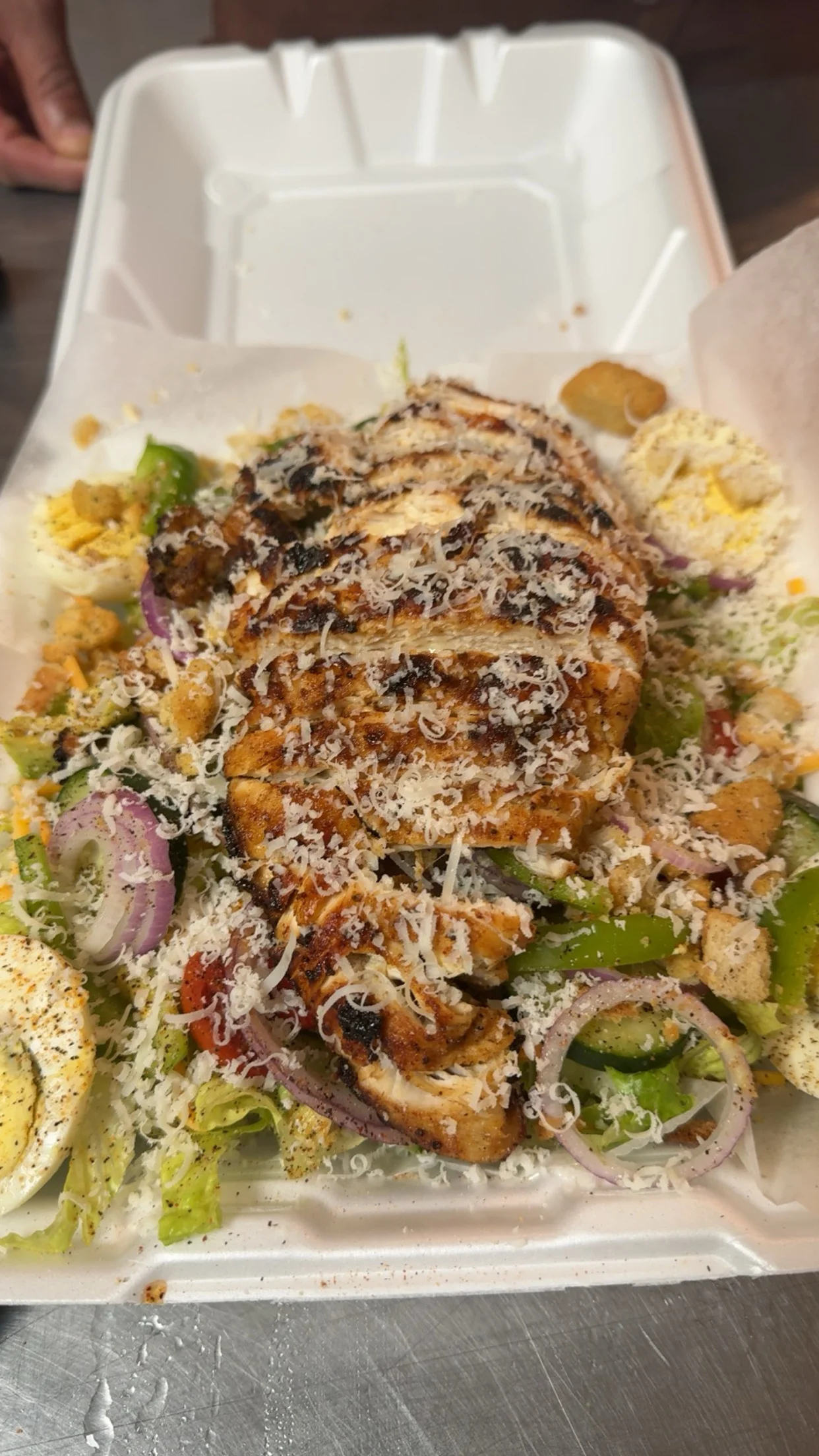 A colorful salad with grilled chicken, fresh vegetables, and grated cheese, served in a takeout container.