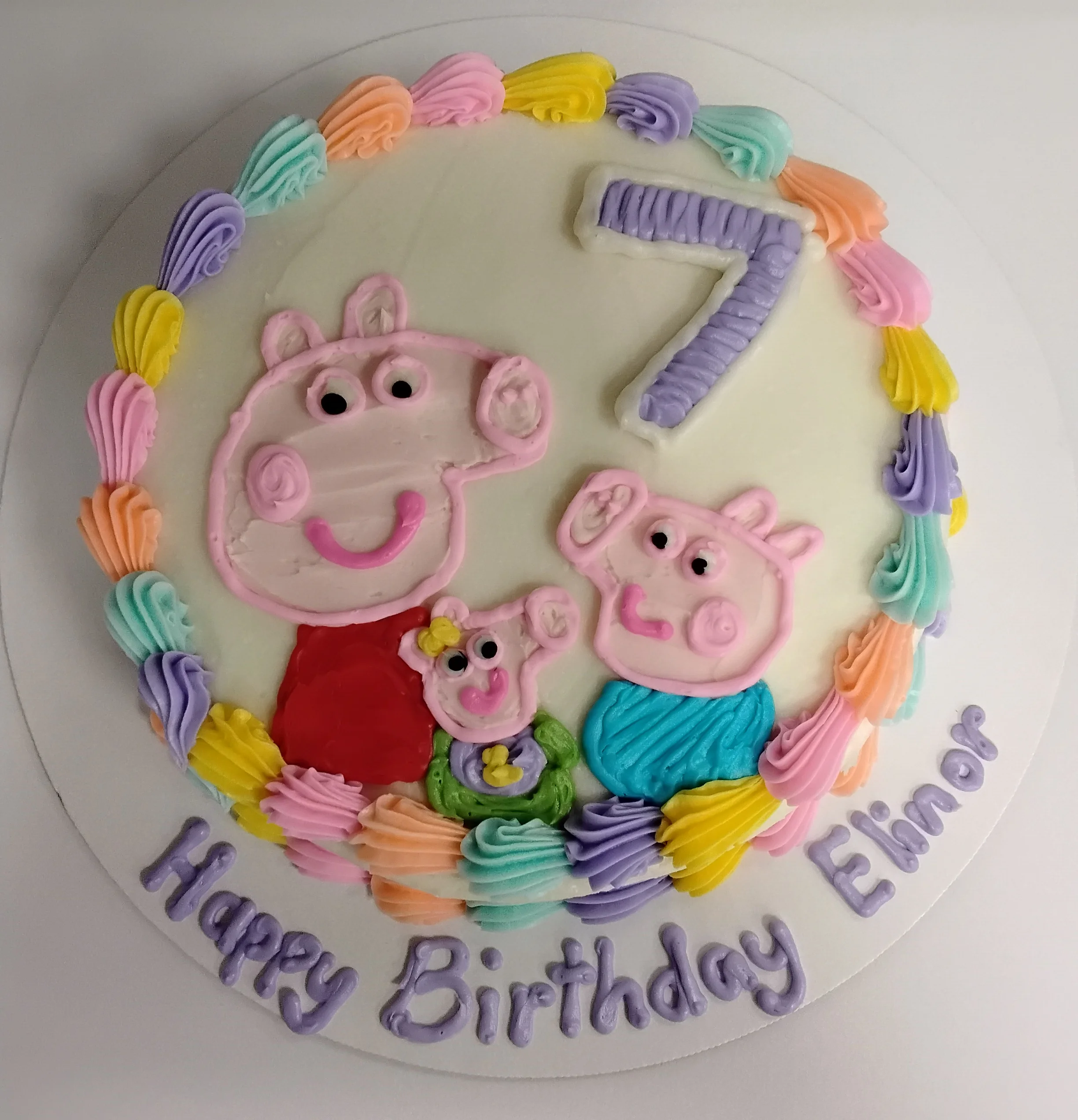 Round cake with a colorful border and Peppa Pig characters drawn in icing with the number 7 above them. Written on the cake board says "Happy Birthday Elinor"