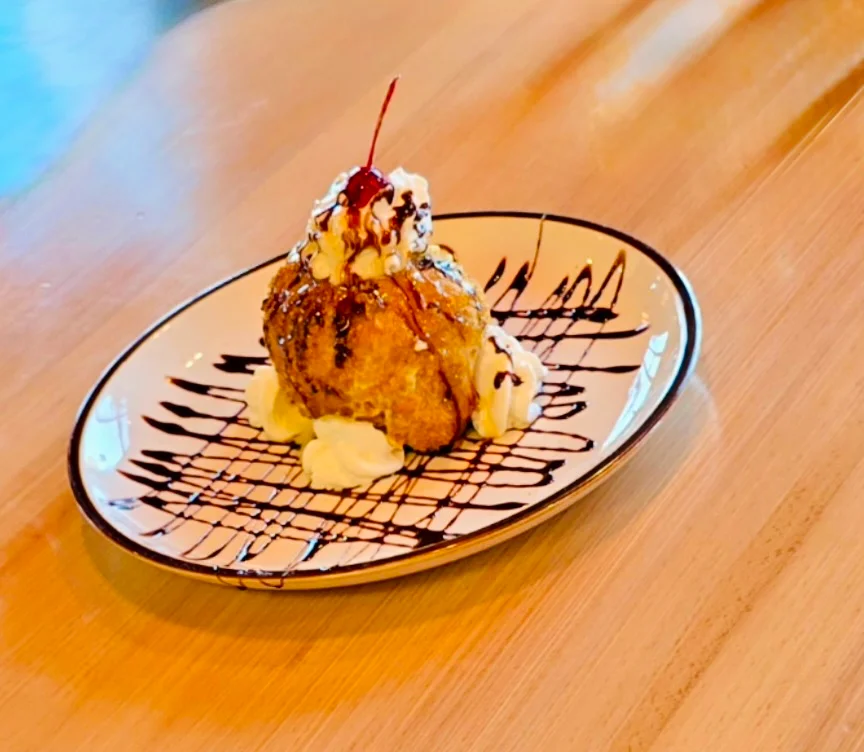 A dessert featuring a fried treat topped with whipped cream, a cherry, and chocolate drizzle, presented on a decorative plate.