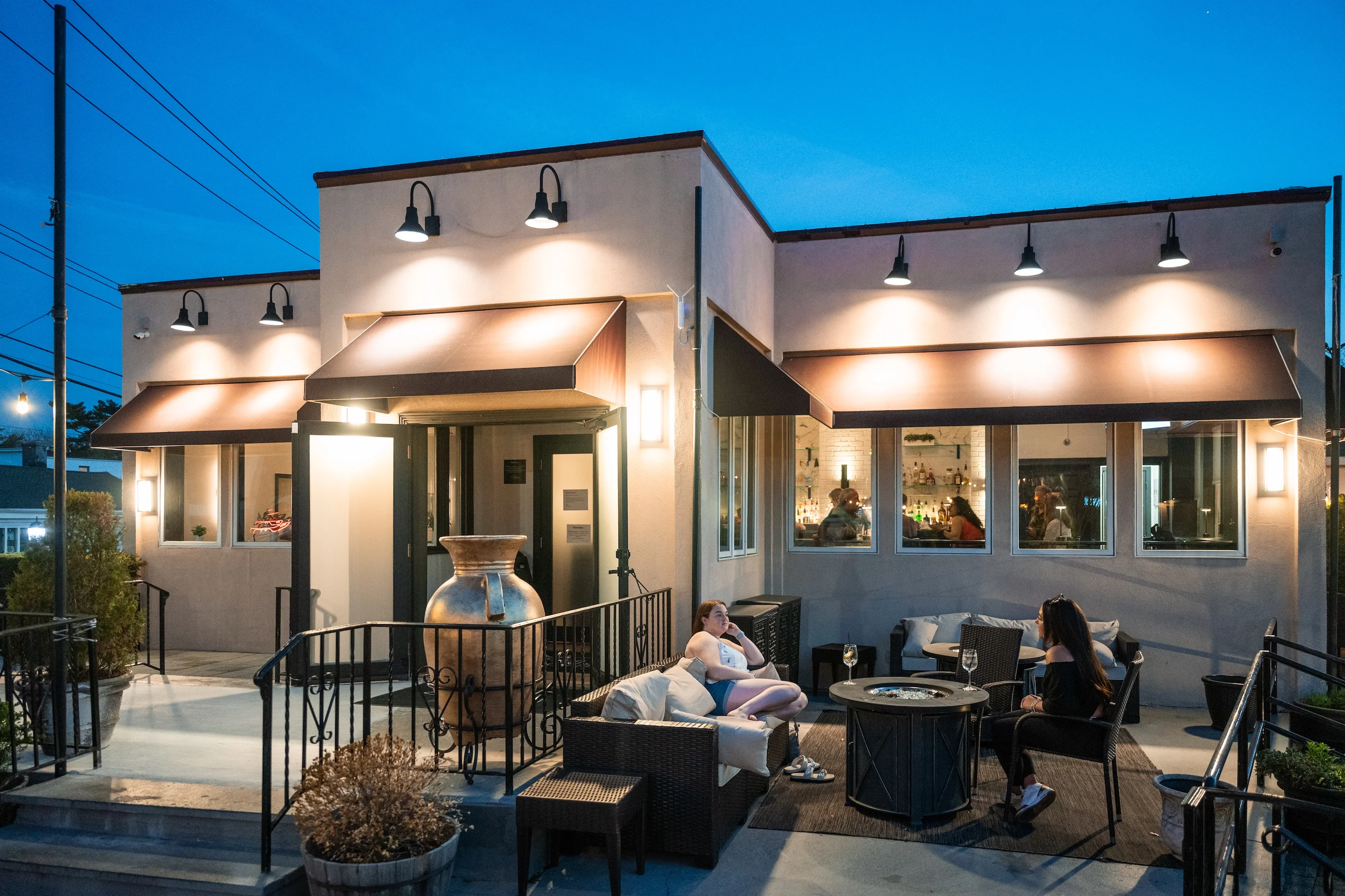 Outdoor patio of Toscana on Main in Eastchester NY at twilight, with warm lighting enhancing a cozy atmosphere. Two women relax on wicker seating, chatting.