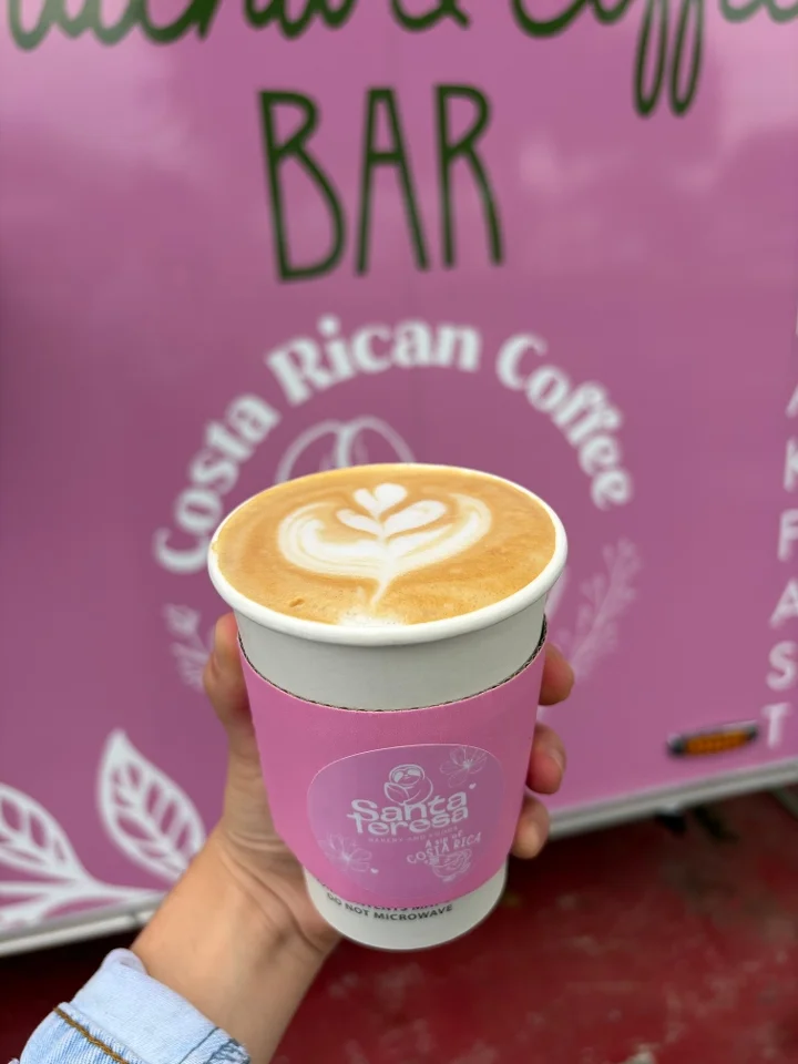 A cup of latte art coffee held in a hand, featuring a beautiful leaf design on the surface, against a pink backdrop.