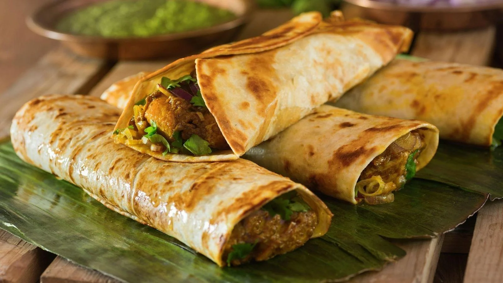 Three rolled flatbreads filled with savory ingredients, resting on a banana leaf, accompanied by a small bowl of green sauce.