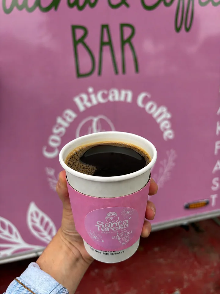 A cup of dark coffee, served in a paper cup, displayed in front of a vibrant pink coffee stand backdrop.