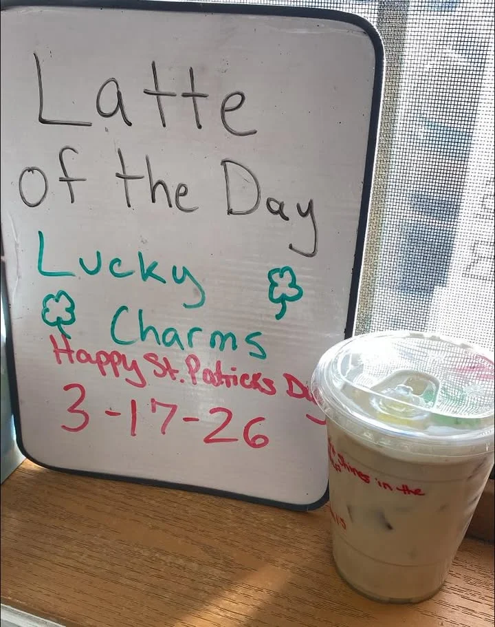 A chalkboard sign announcing the latte of the day, decorated with a promotional message and a cup of iced coffee beside it.
