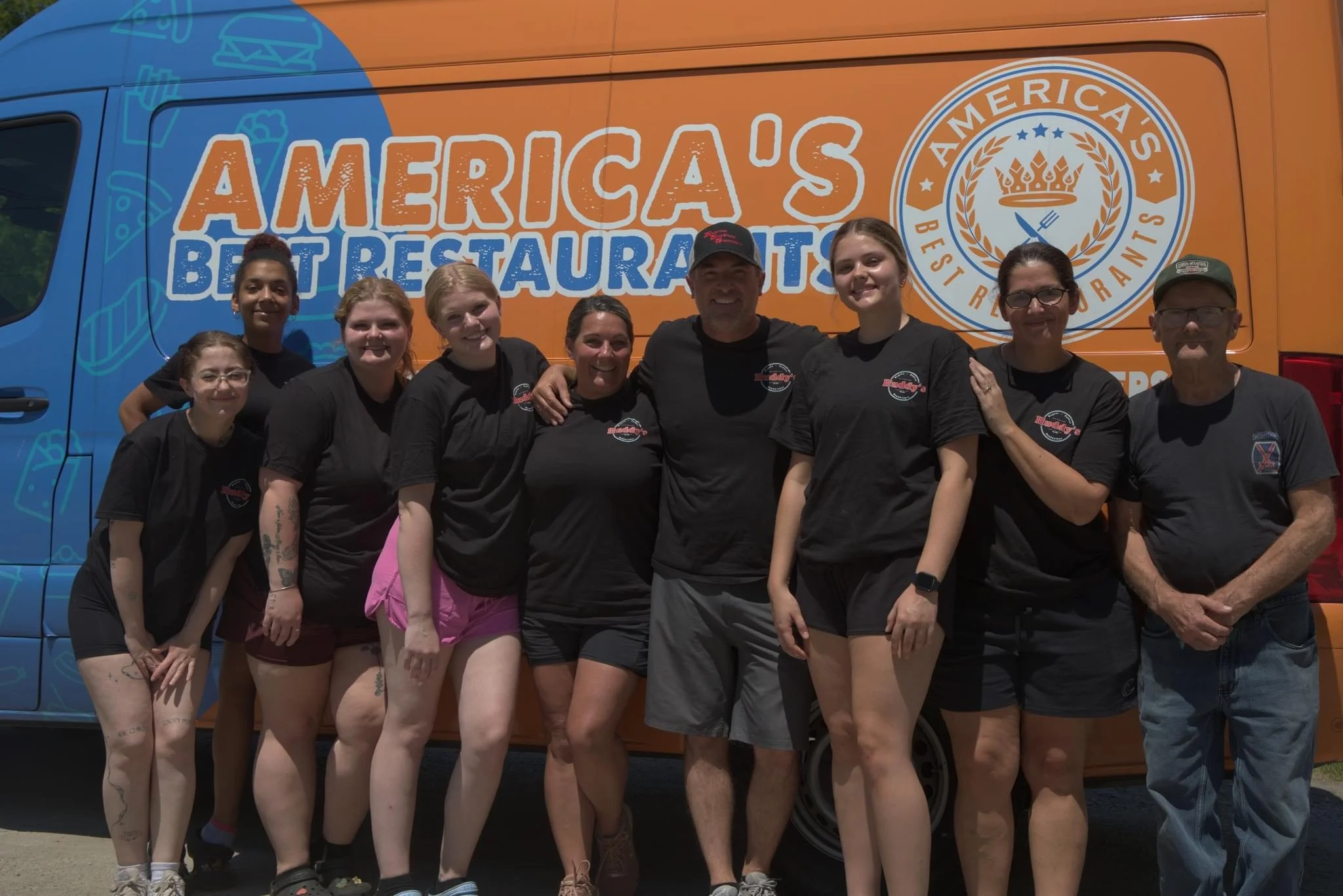 A group of employees posing in front of a brightly colored food truck featuring 'America's Best Restaurants' branding.