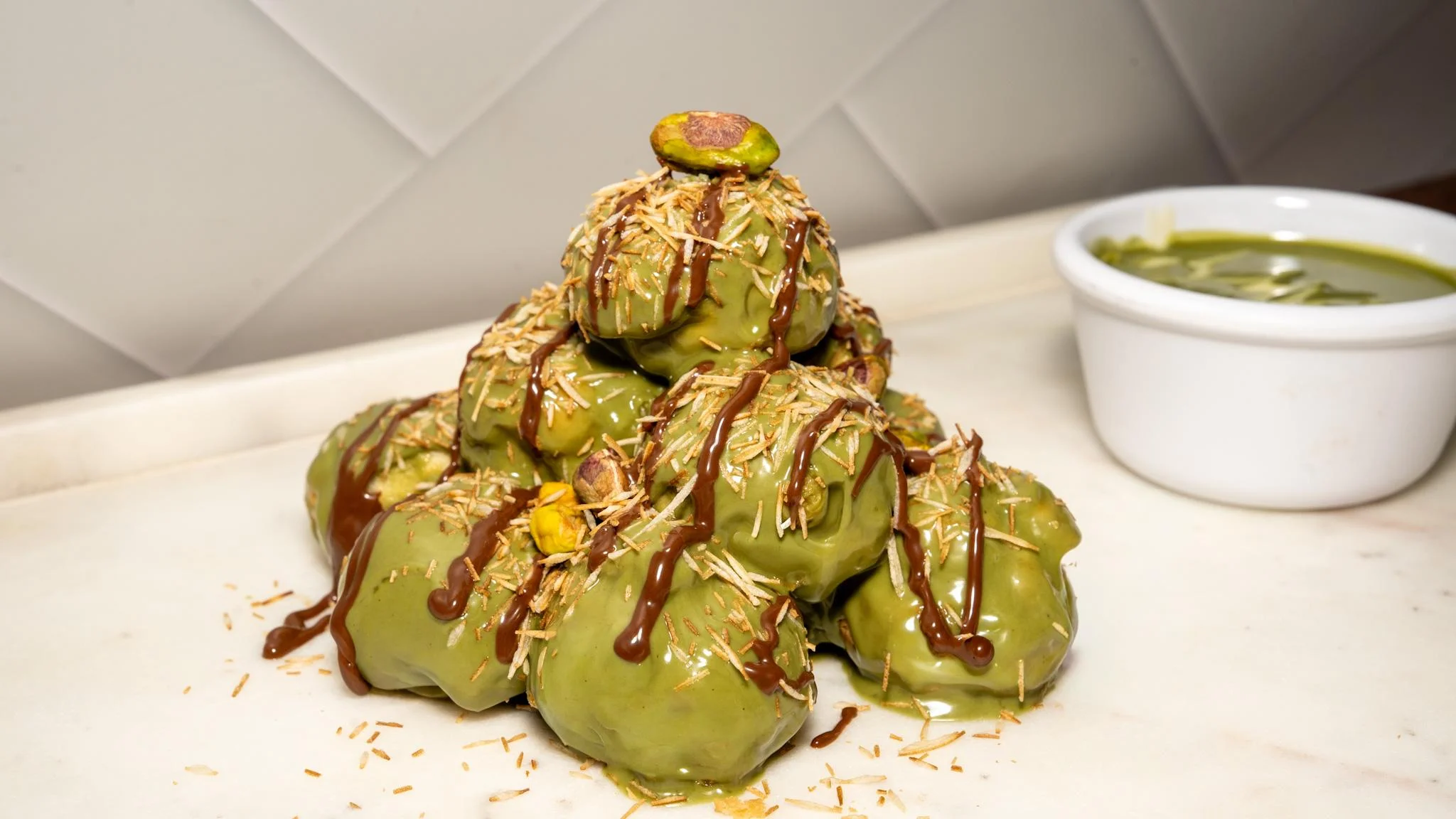 A mound of green sweets topped with a pistachio and drizzled with chocolate, garnished with shredded coconut.