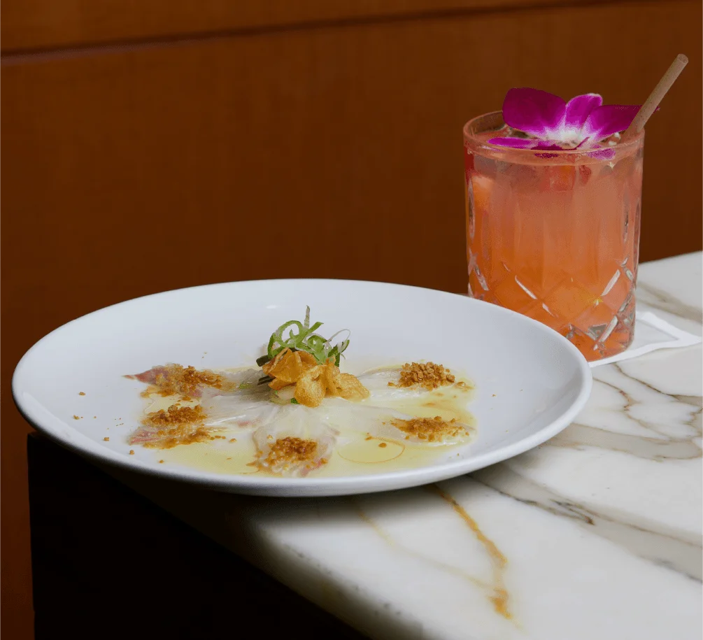 A white plate containing a fan of thinly sliced fish carpaccio dressed with olive oil, microgreens, and toasted breadcrumbs, sitting on the corner of a white marbled table next to a pink cocktail garnished with a purple orchid flower.