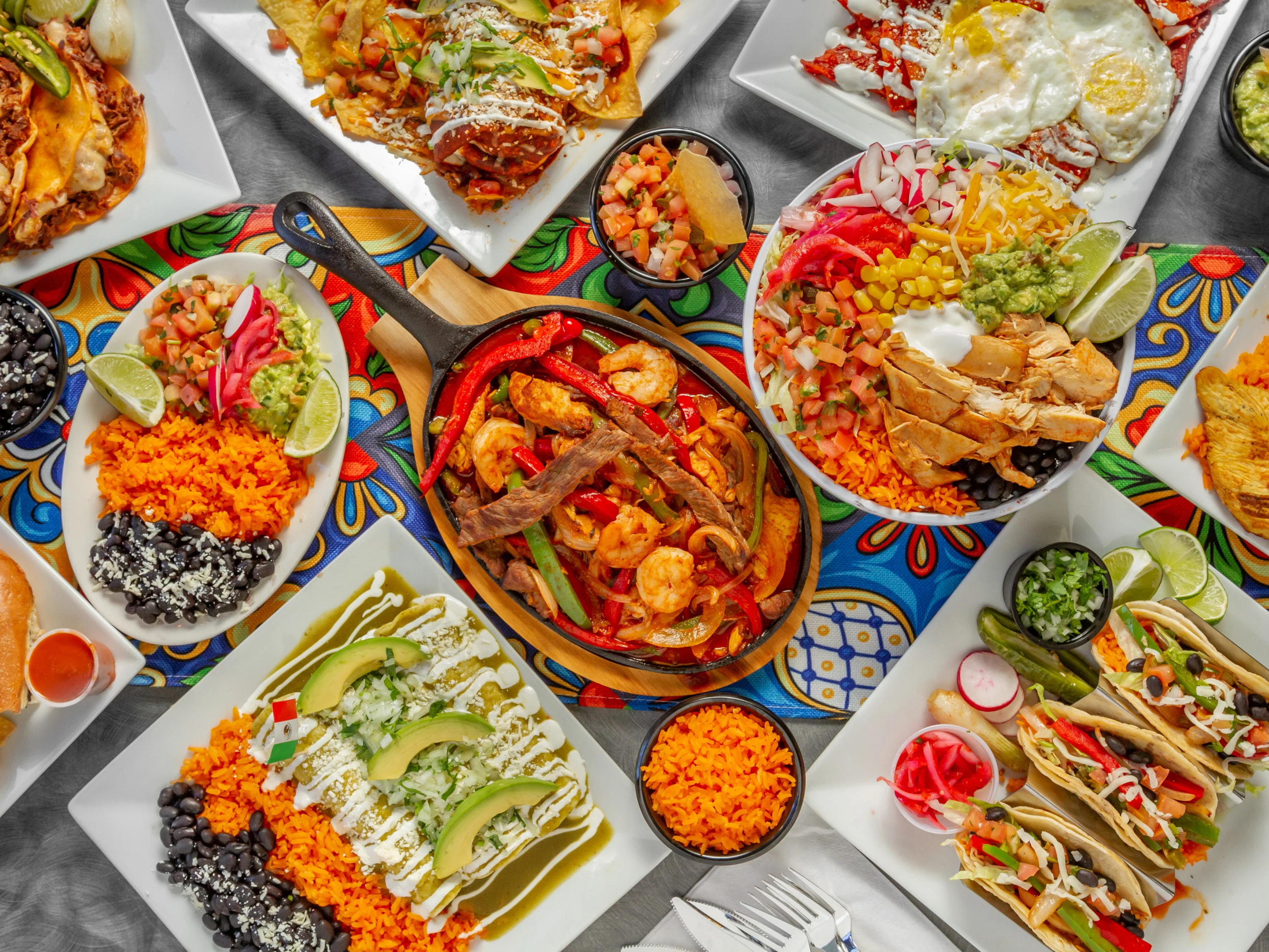 A vibrant spread of Mexican dishes including fajitas, tacos, and a variety of sides arranged on a colorful tablecloth.