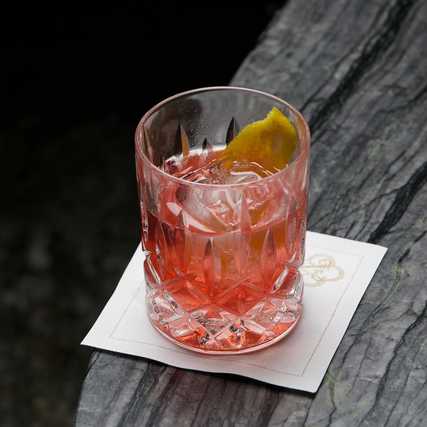 A vibrant red cocktail with a large ice cube and a citrus peel garnish, served in a crystal cut rocks glass on a white square napkin over a dark, textured stone surface.