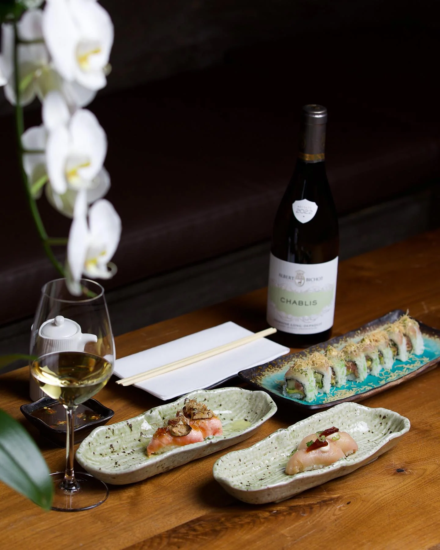 An elegant table setting featuring a bottle of Albert Bichot Chablis wine next to a glass of white wine, a colorful sushi roll on a teal platter, and two individual pieces of nigiri sushi served on light green ceramic dishes, accented by white orchid blossoms.