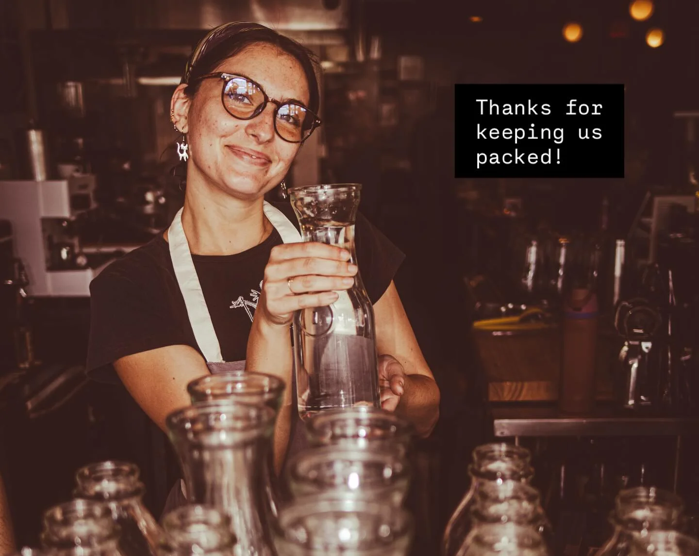 A worker in a restaurant smiling while holding a glass pitcher among a collection of empty glass bottles.