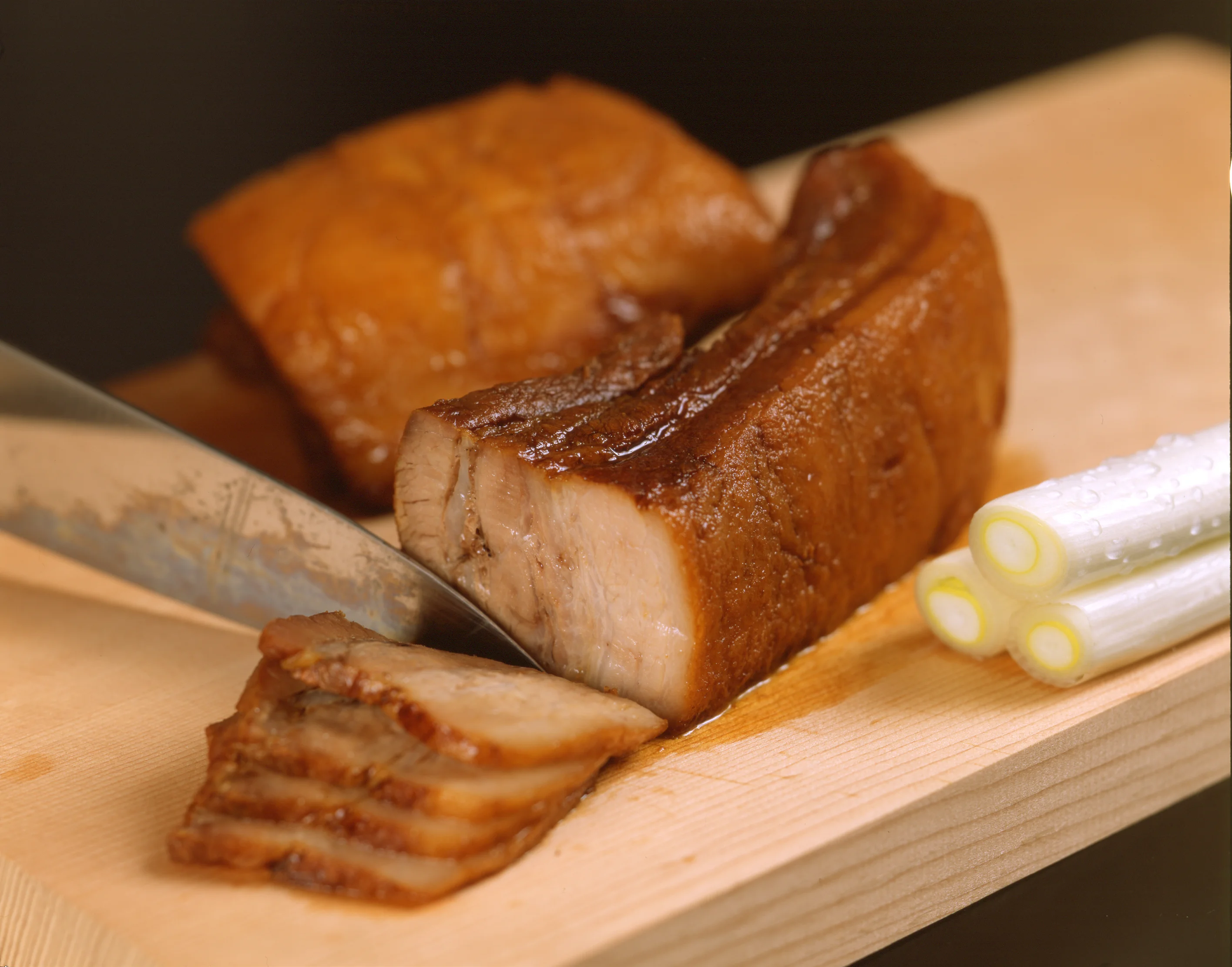 Chashu pork belly being sliced on a wooden cutting board next to green onion stalks