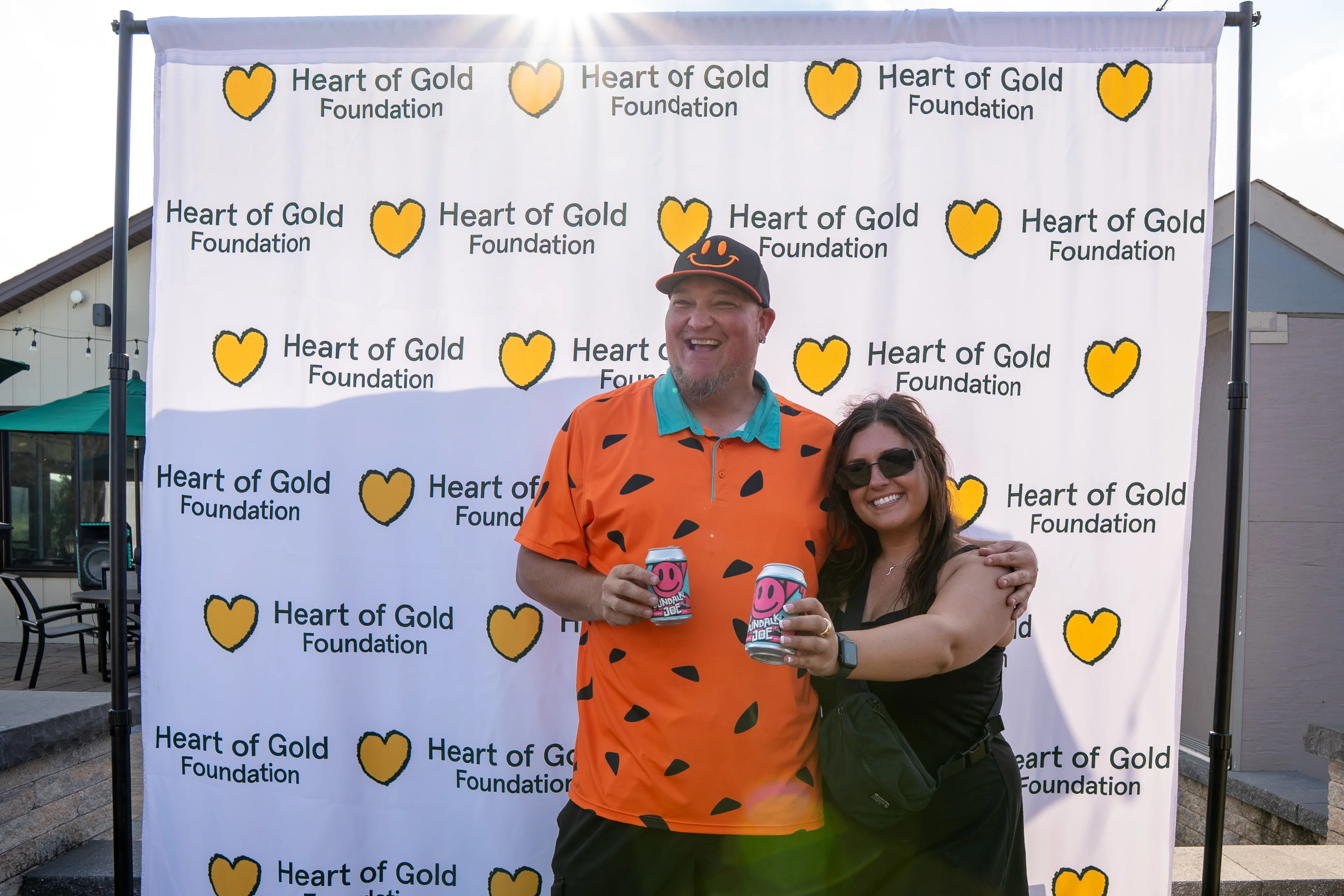 Two people smiling with drinks in front of a Heart of Gold Foundation backdrop.
