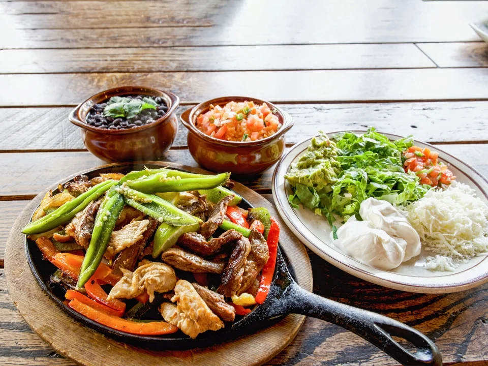 A sizzling platter filled with grilled meats and bell peppers, served with sides of black beans, fresh pico de gallo, guacamole, lettuce, and sour cream on a wooden table.