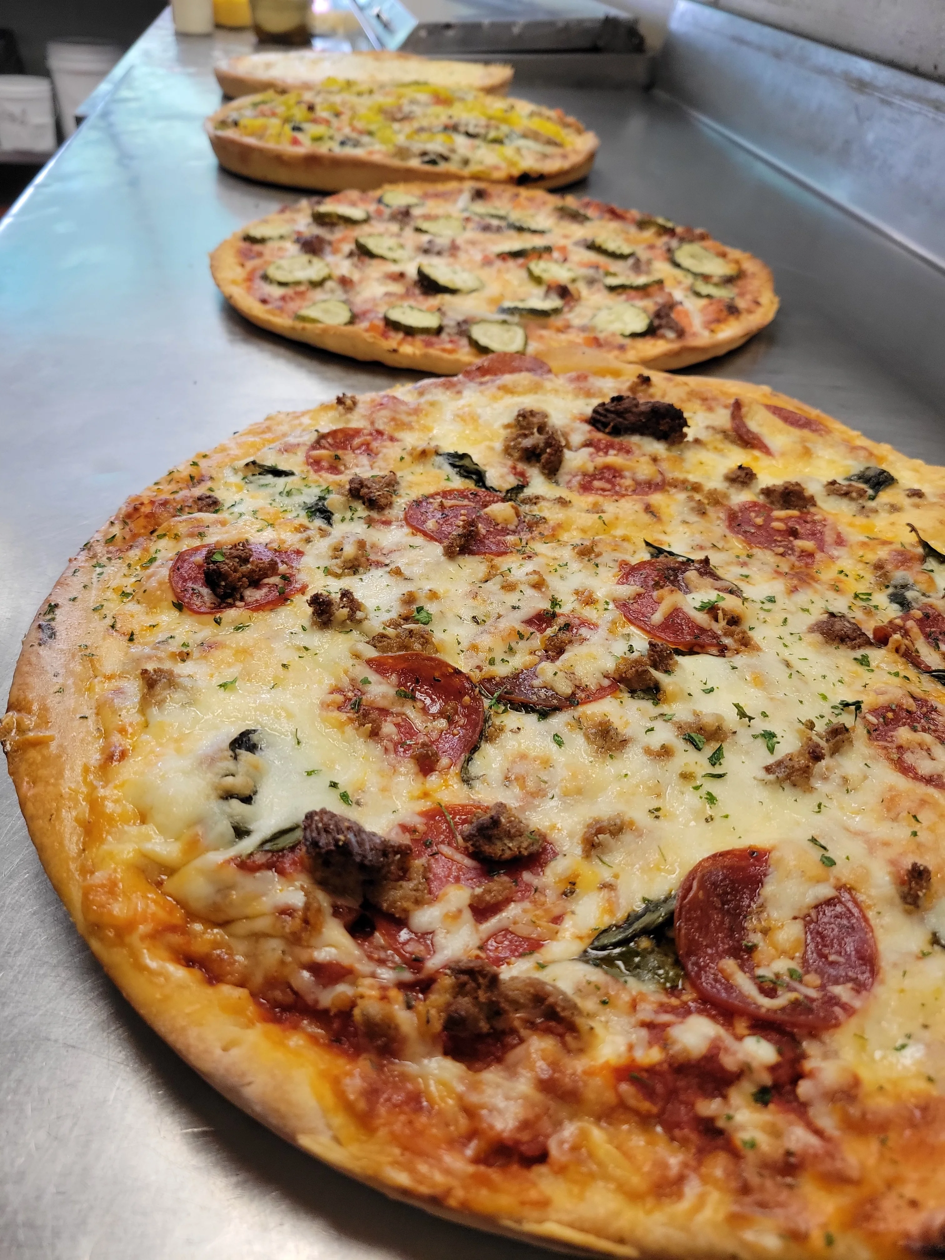 Three freshly baked pizzas placed on a countertop. The front pizza is topped with pepperoni and ground meat, while the other two feature vegetables and different toppings.