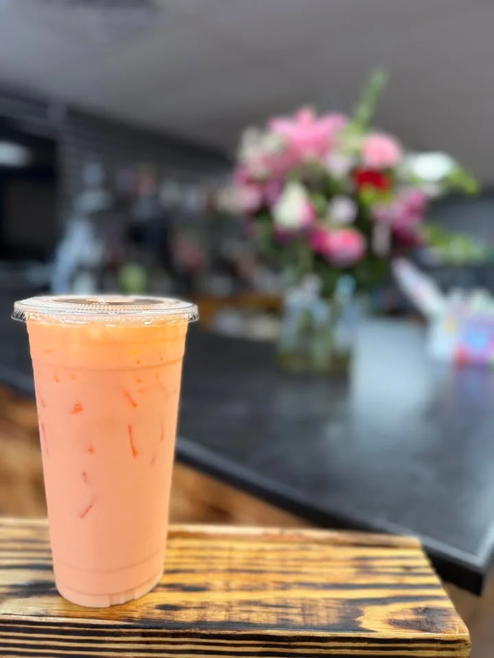 A tall cup of light pink drink filled with ice, placed on a wooden surface, with a floral arrangement in the background.