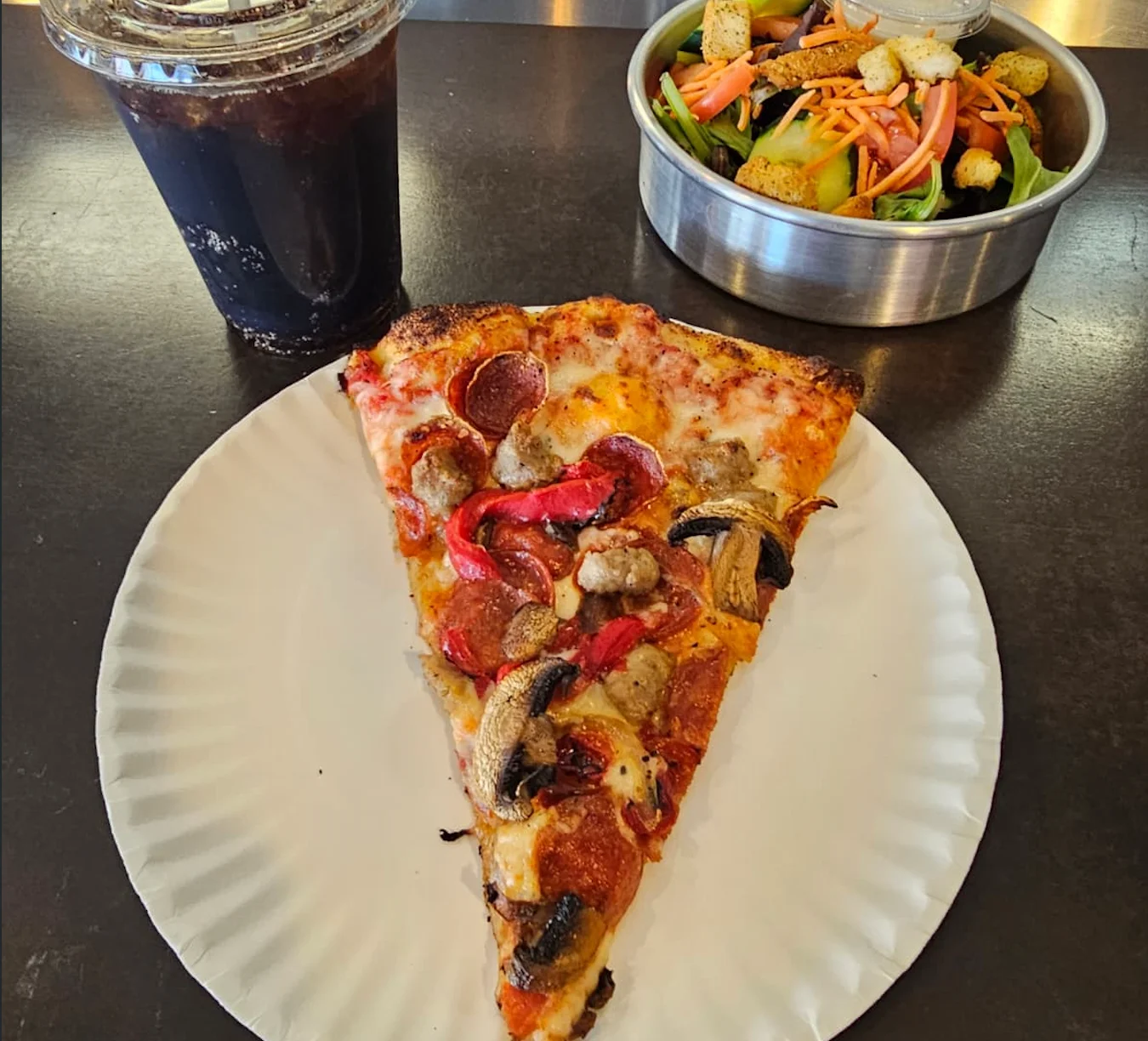 New York-style pizza slice with pepperoni, sausage, vegetables, salad, and cola