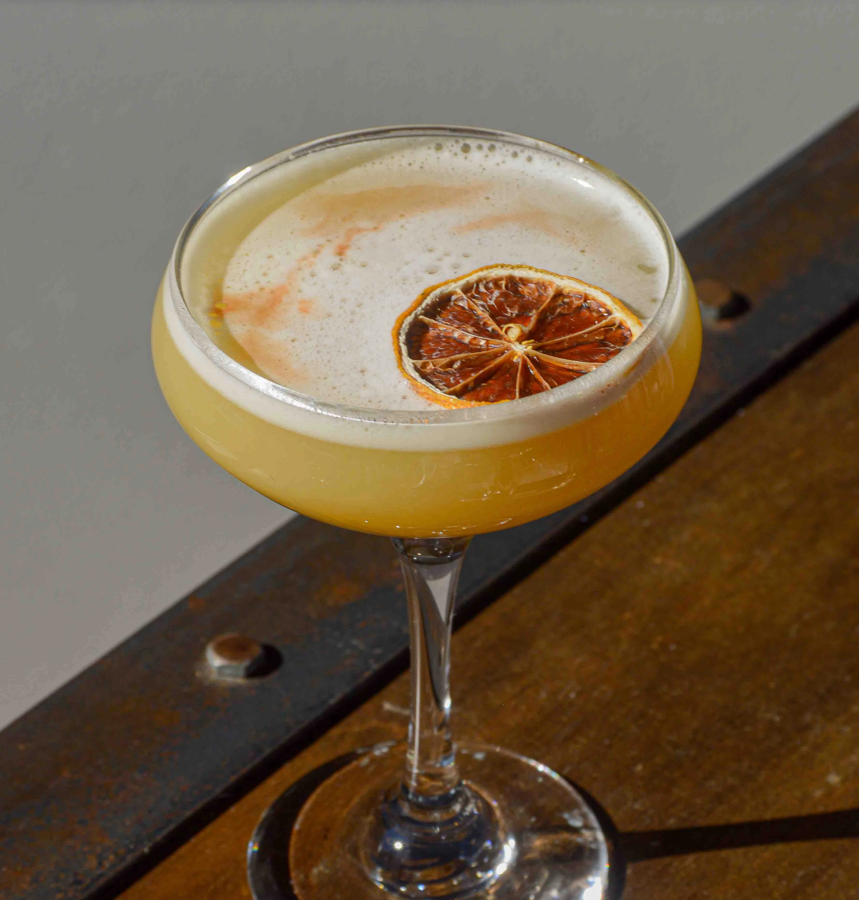 A dried lemon slice sits atop a pineapple sour on a wooden table
