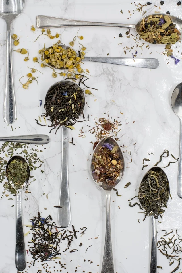Variety of loose teas and herbs in silver spoons on marble surface