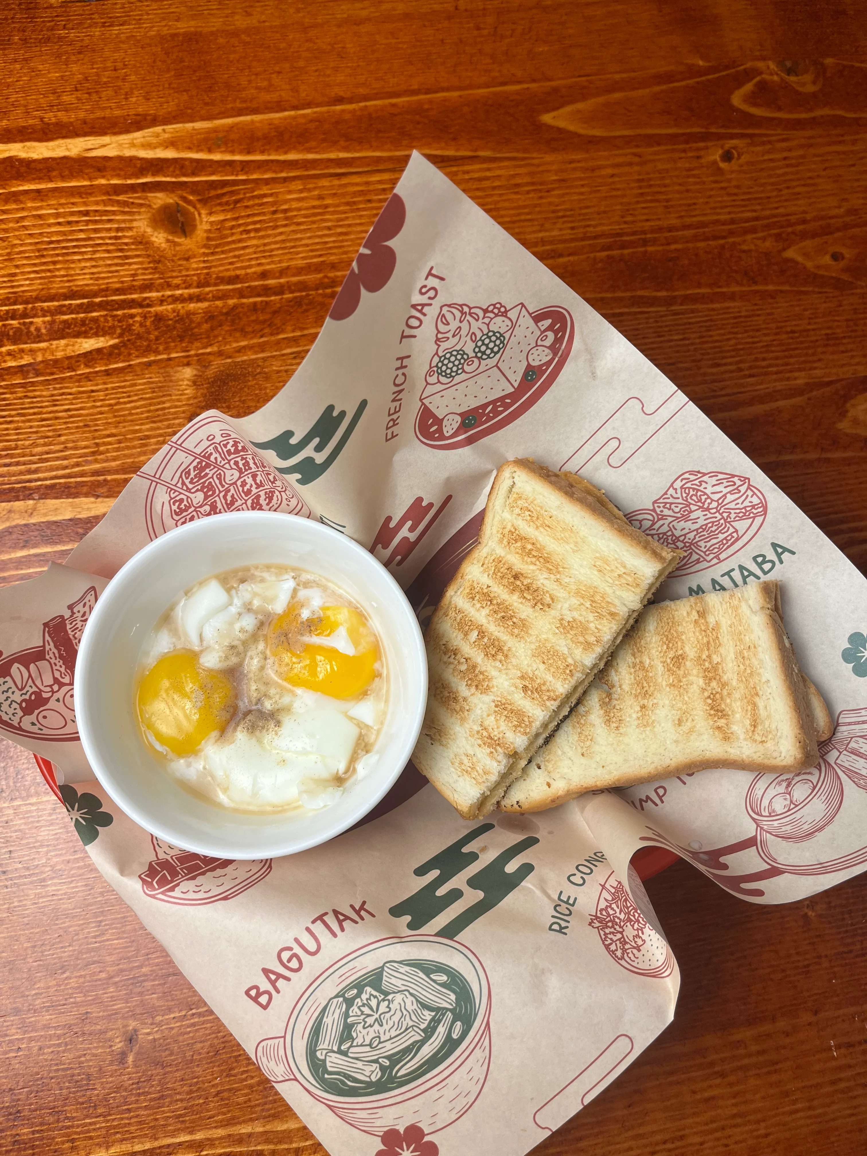 A serving of soft-boiled eggs with two egg yolks in a bowl alongside slices of toasted bread. The dish is placed on a decorative paper featuring various food illustrations.