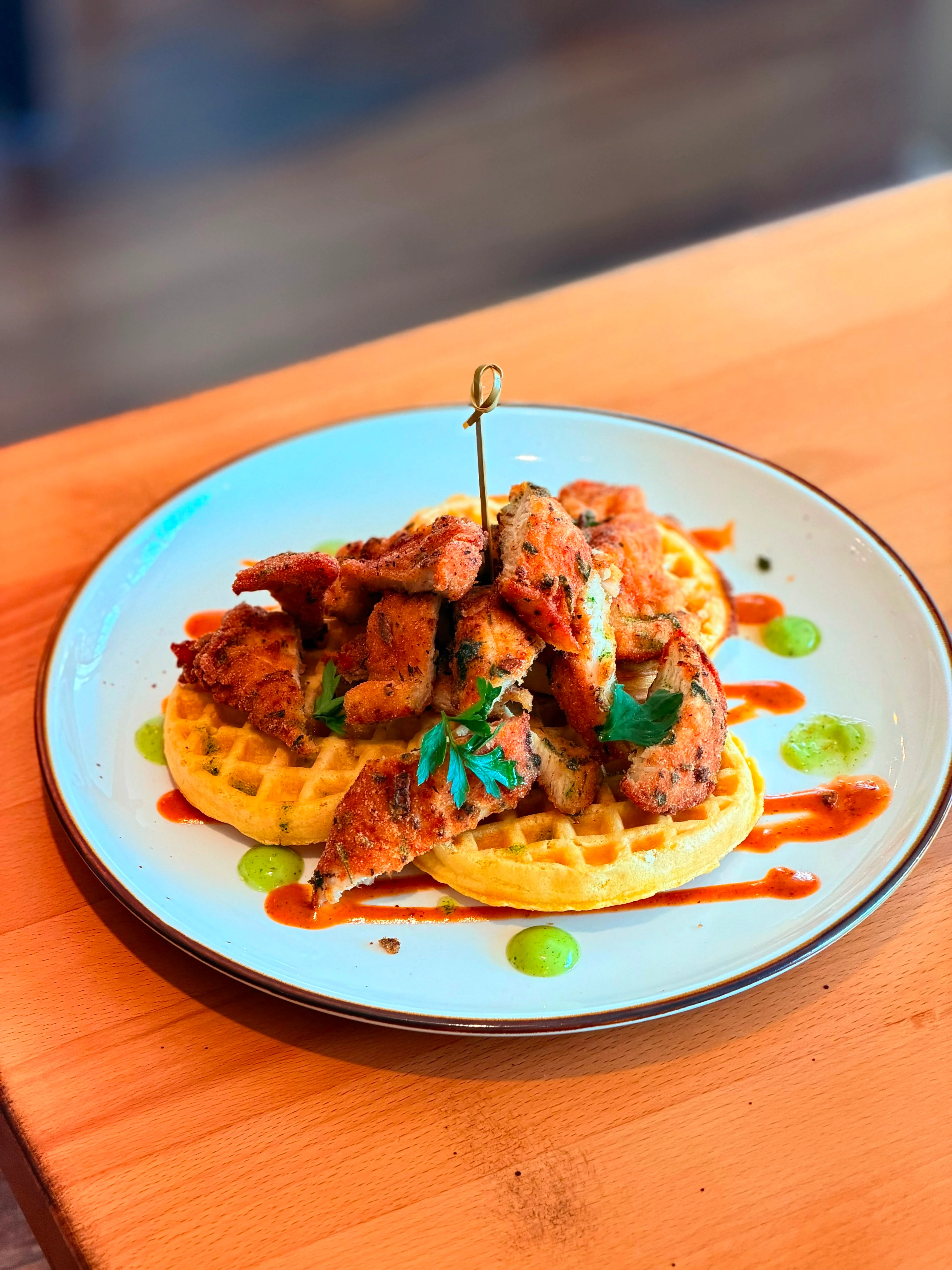 A plate of crispy chicken served on waffles, garnished with a green sauce and a drizzle of spicy sauce.