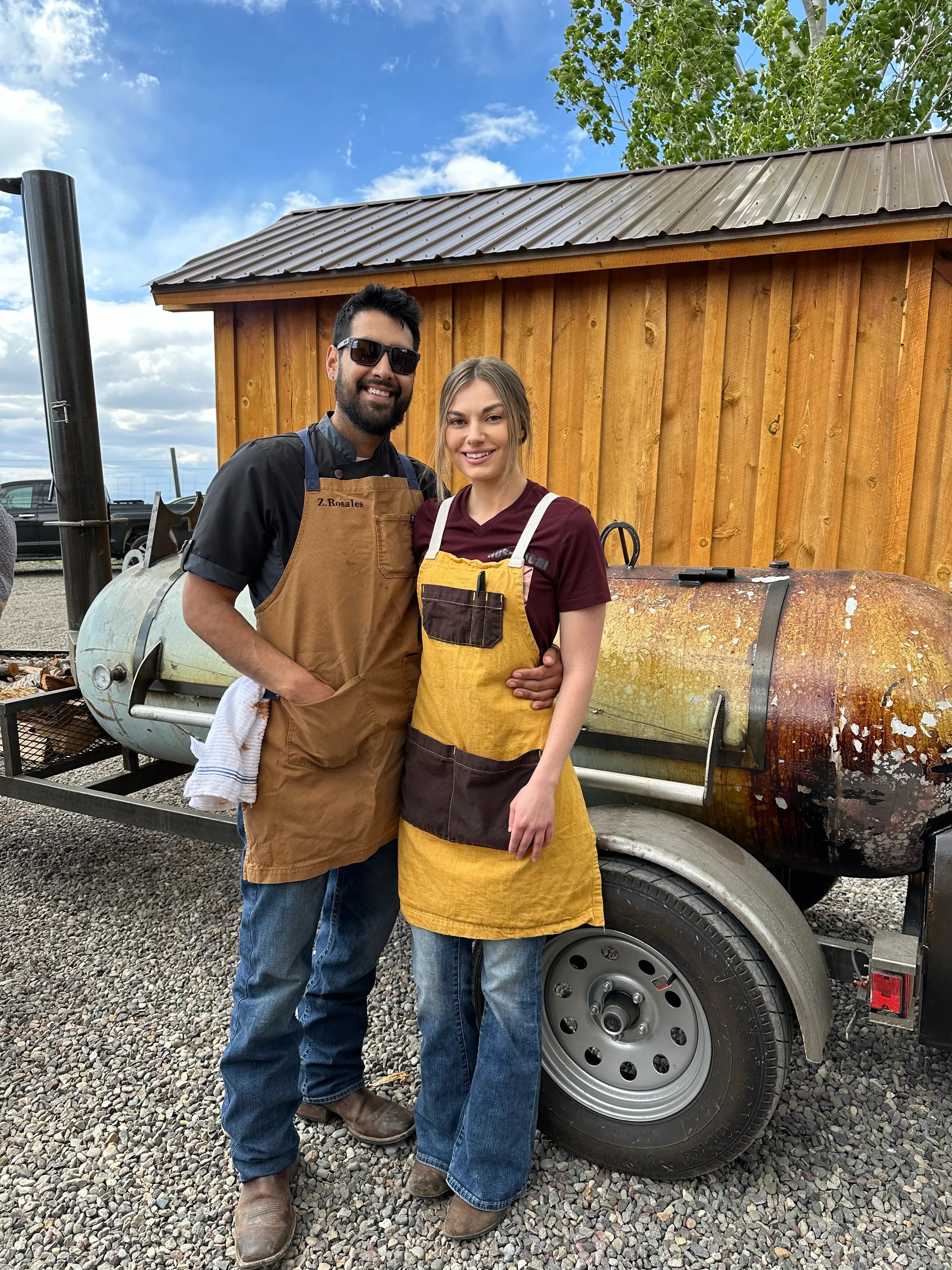 Two chefs posing in front of a smoker trailer outdoors