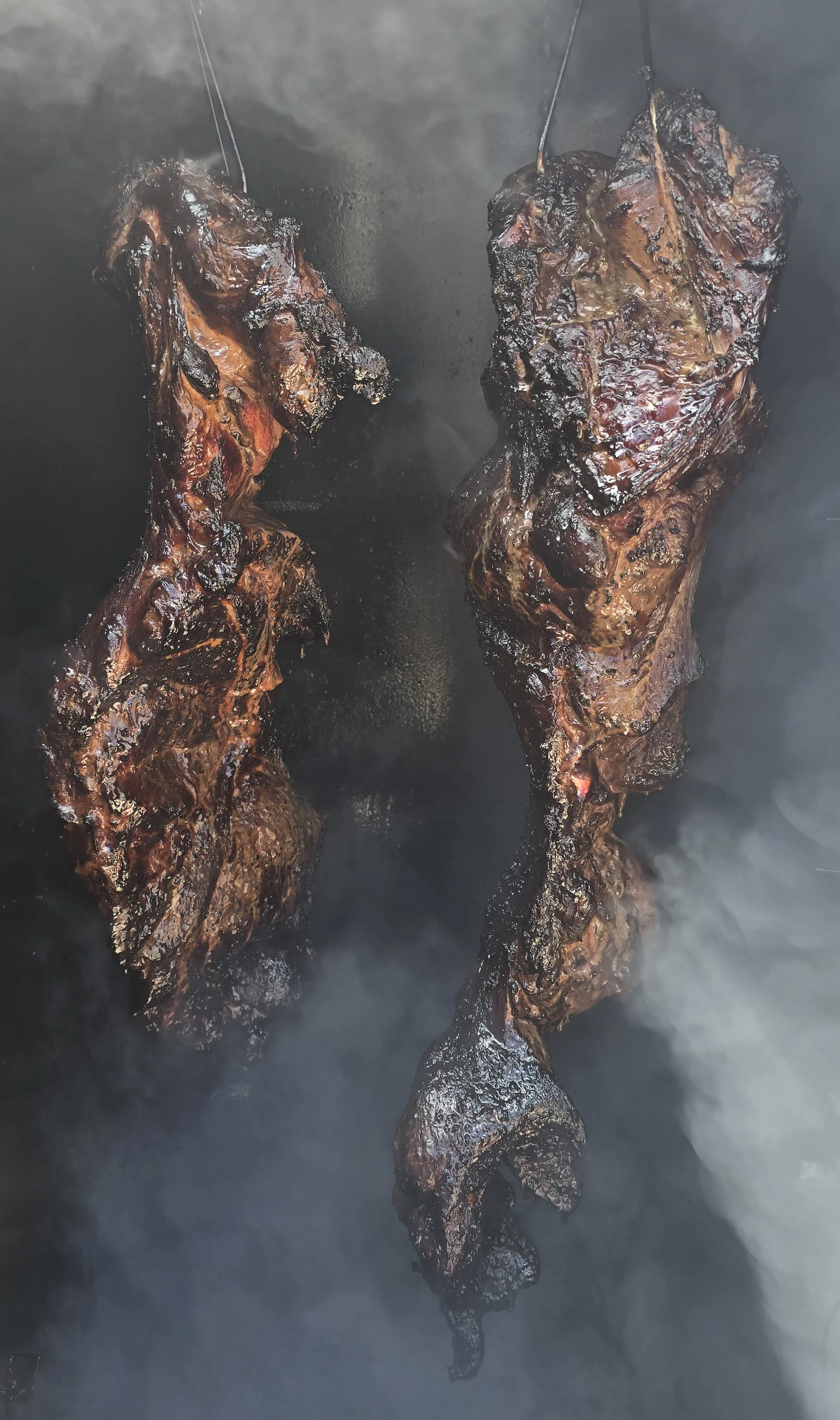 Two large pieces of smoked meat hanging in a smoky environment.