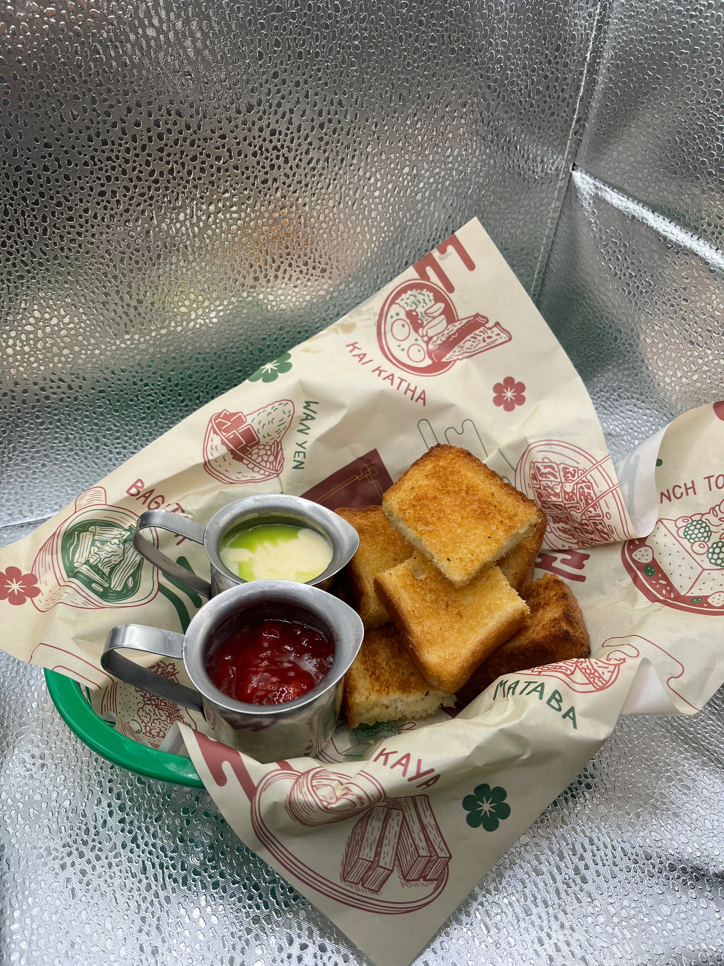 A plate of toasted bread squares served with two small containers of sauces, on patterned paper.