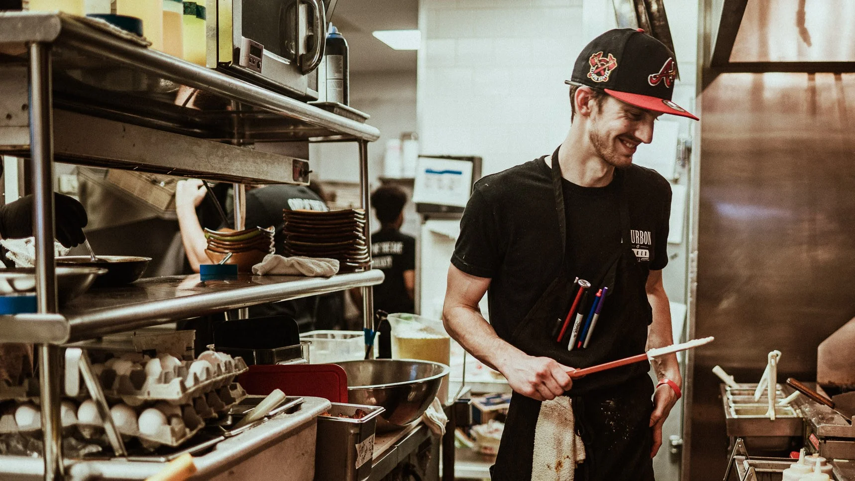 Chef smiling while preparing food in a busy restaurant kitchen.