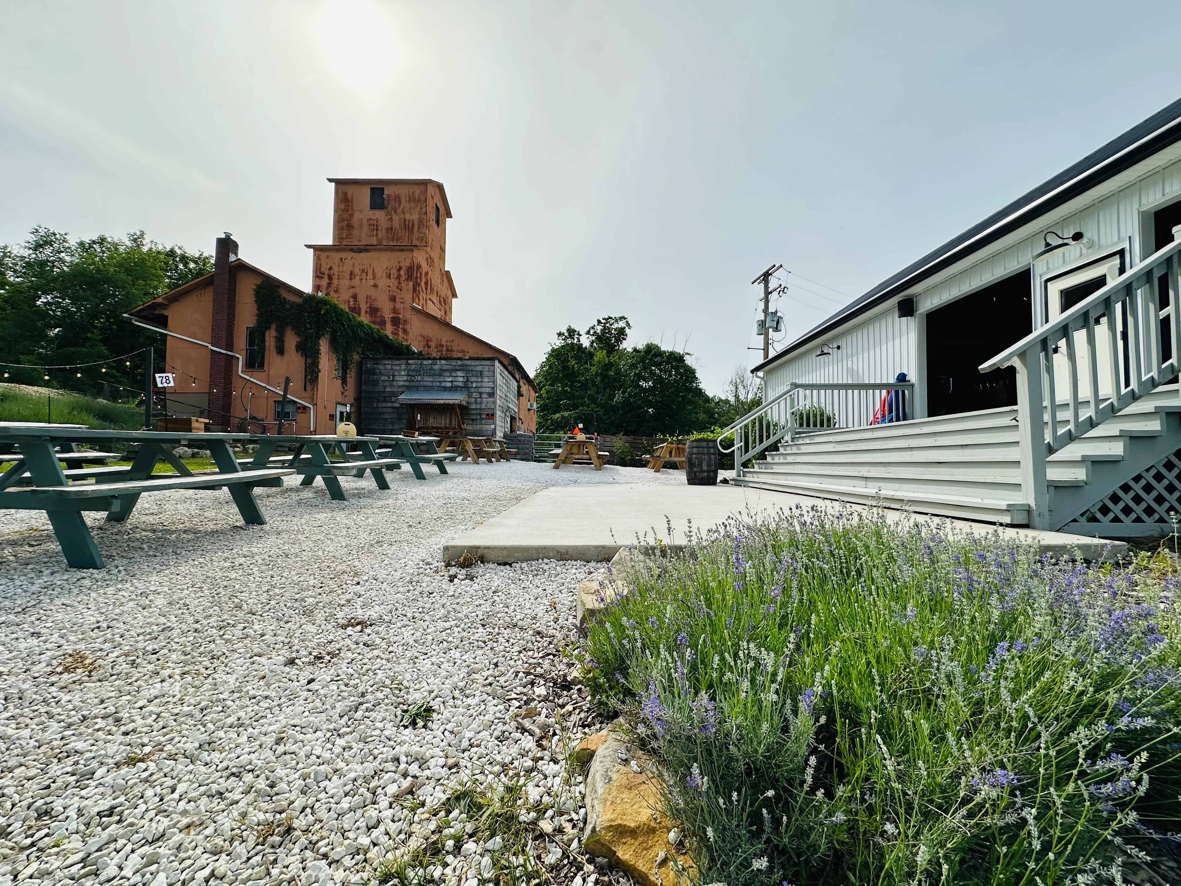 An outdoor seating area with wooden picnic tables, set against a rustic backdrop of an orange building and greenery under a sunny sky.