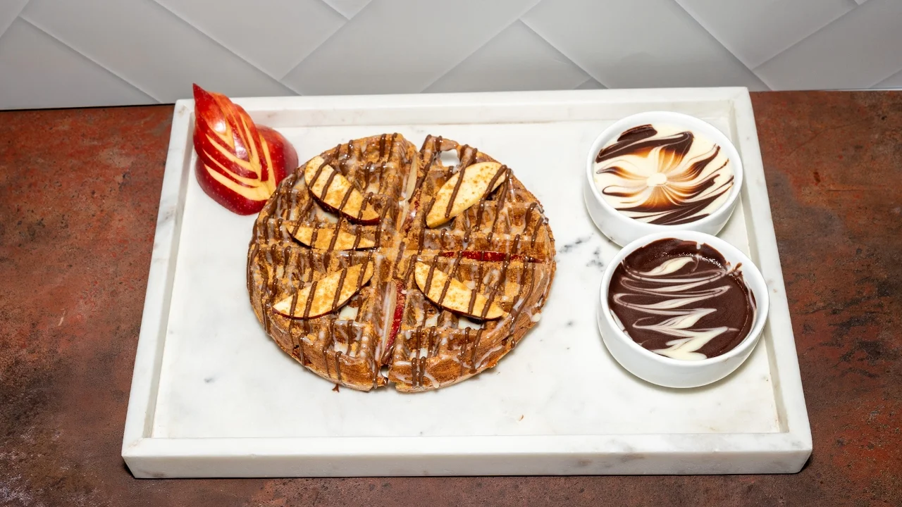 A round dessert topped with apple slices, drizzled with caramel, accompanied by two bowls of chocolate and creamy sauce.