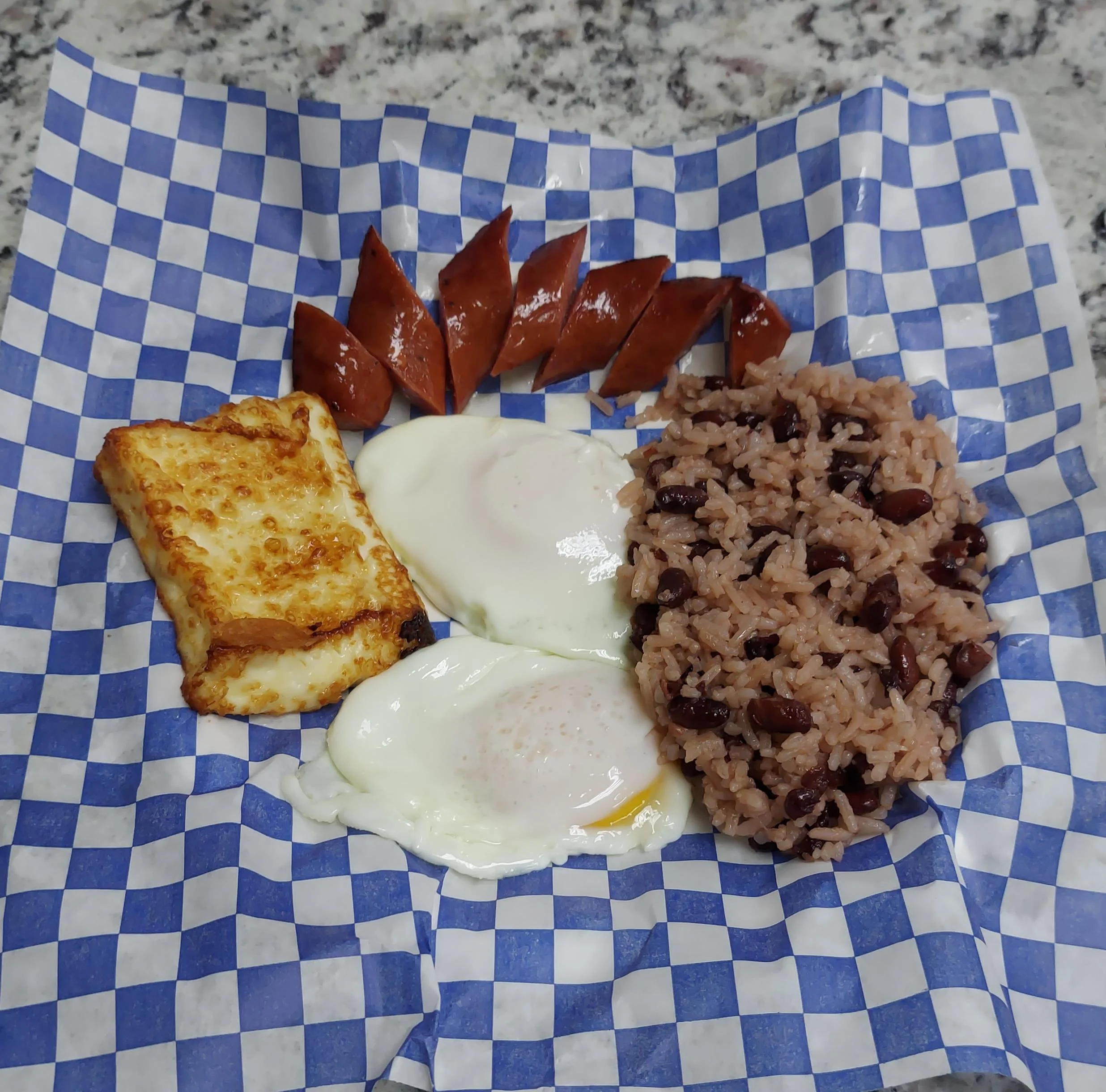 A portion of rice and beans served with two sunny-side-up eggs, slices of sausage, and a piece of fried cheese, presented on checkered paper.