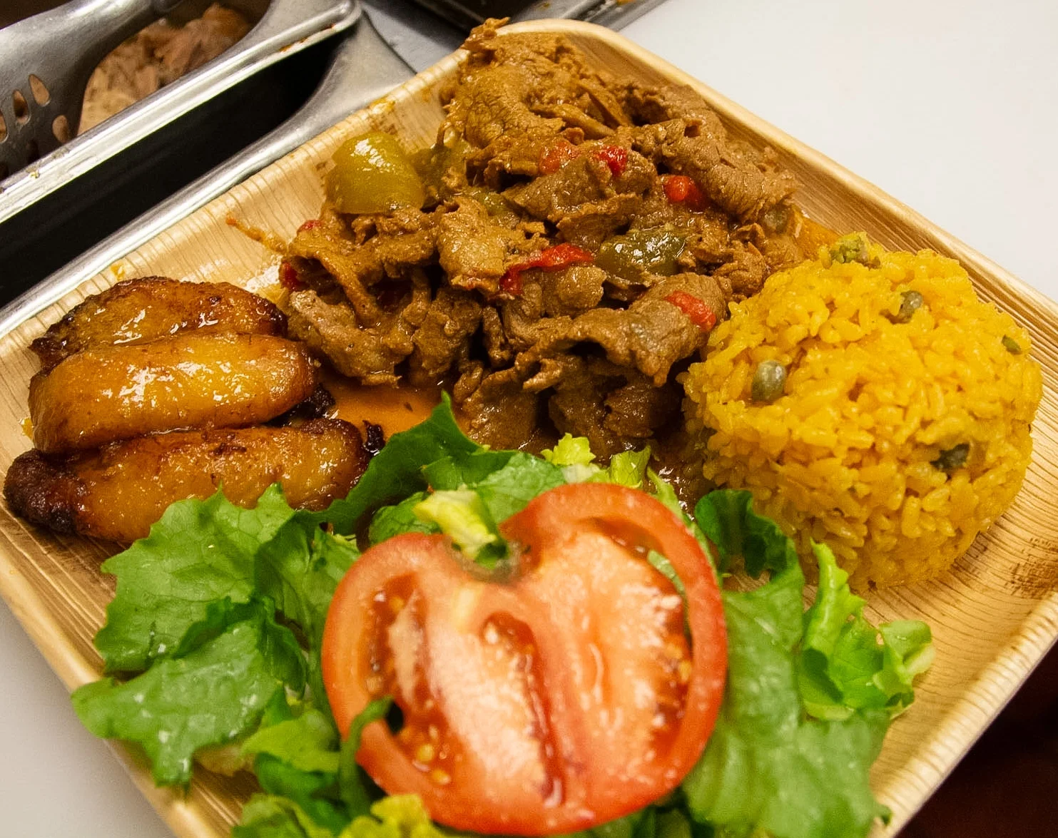 A serving of Puerto Rican food featuring seasoned meat, yellow rice, fried plantains, fresh lettuce, and tomato on a bamboo plate.