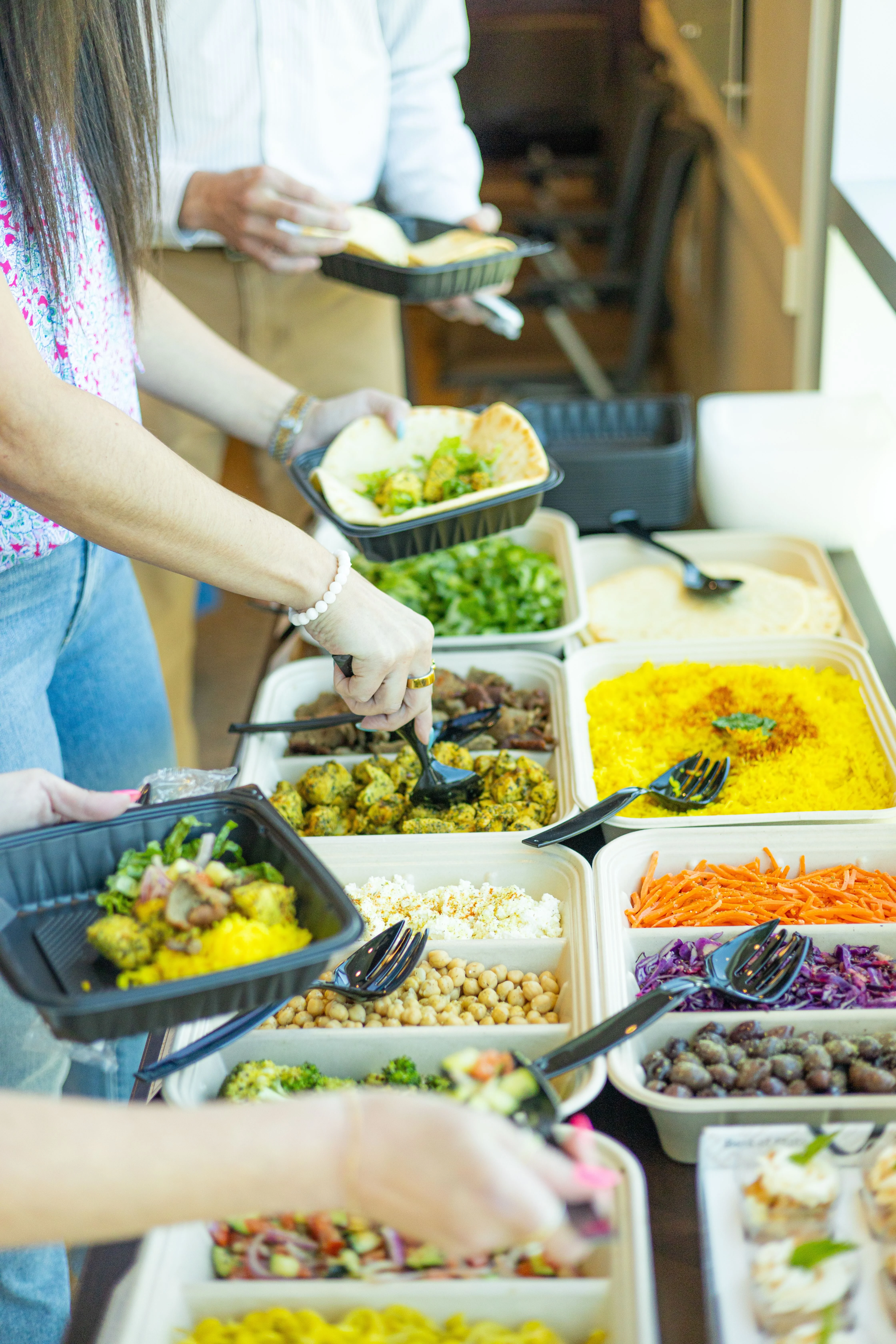 Catering buffet trays with fresh toppings, vegetables, and hummus being served for a build your own buffet at Santorini's Greek Grill.