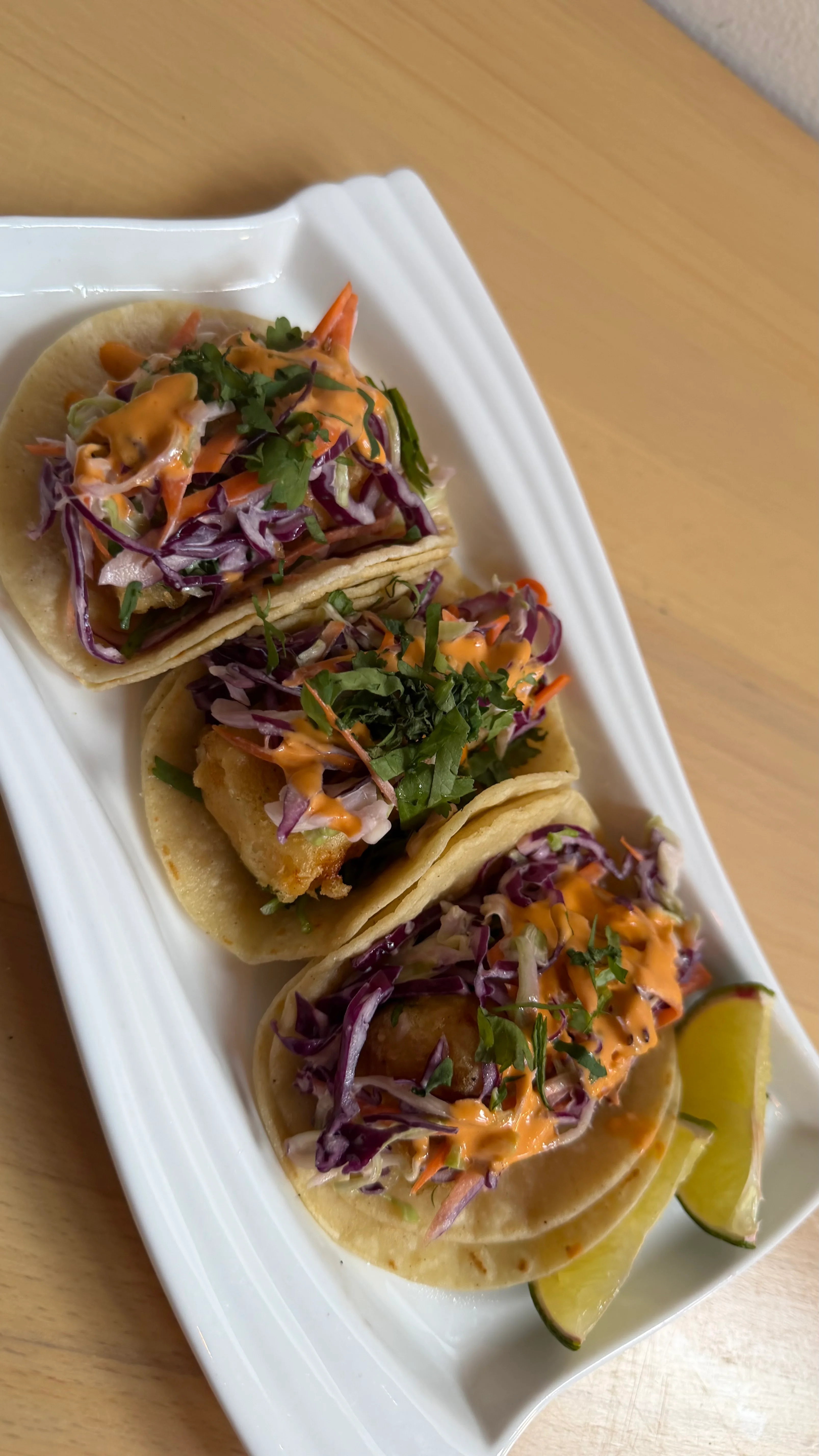 Three tacos filled with cabbage, sauce, and a protein, garnished with cilantro and served with lime wedges on a white plate.