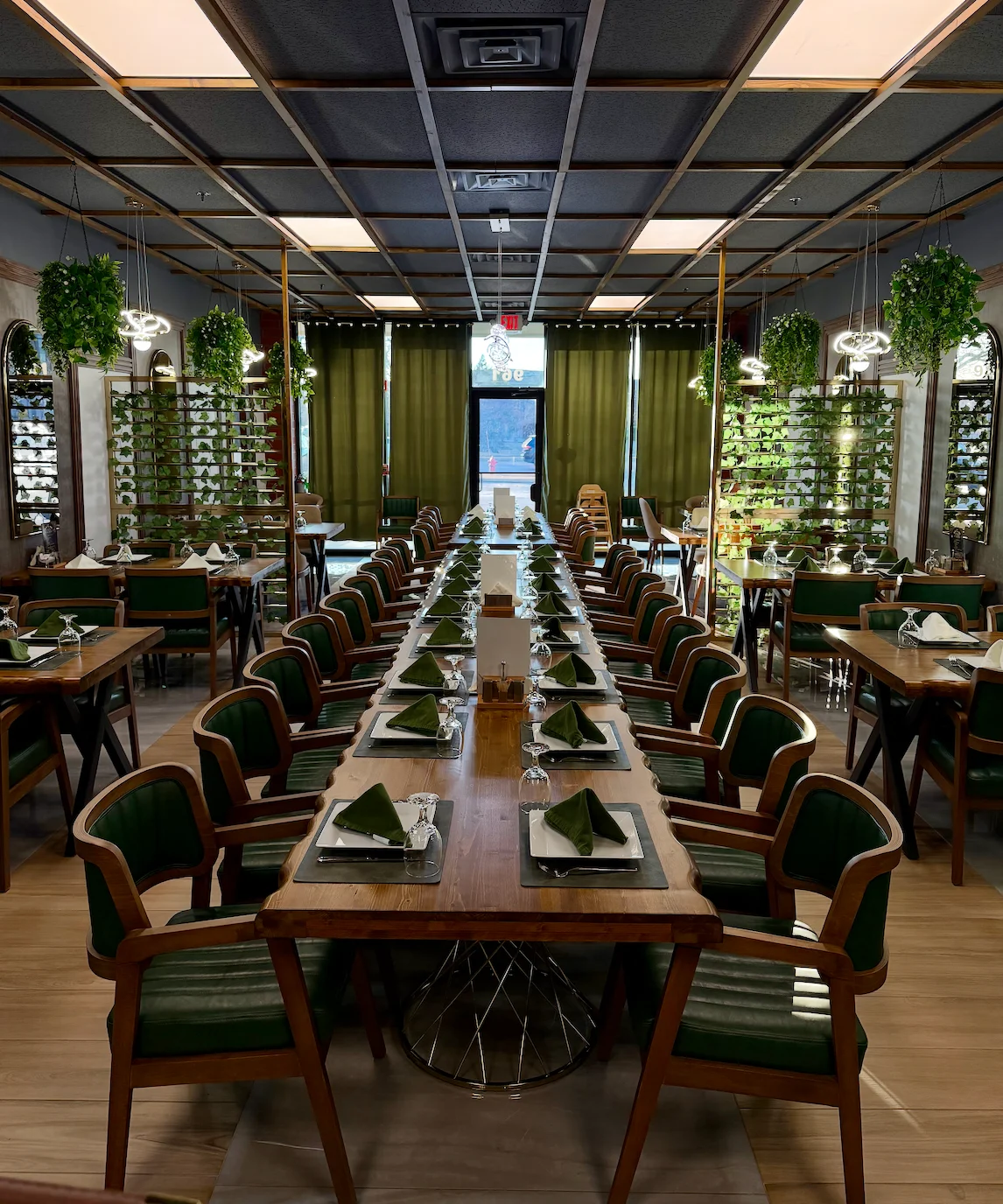 A spacious dining area featuring a long table with green chairs, set with napkins and glassware. The room is adorned with plants and large windows.