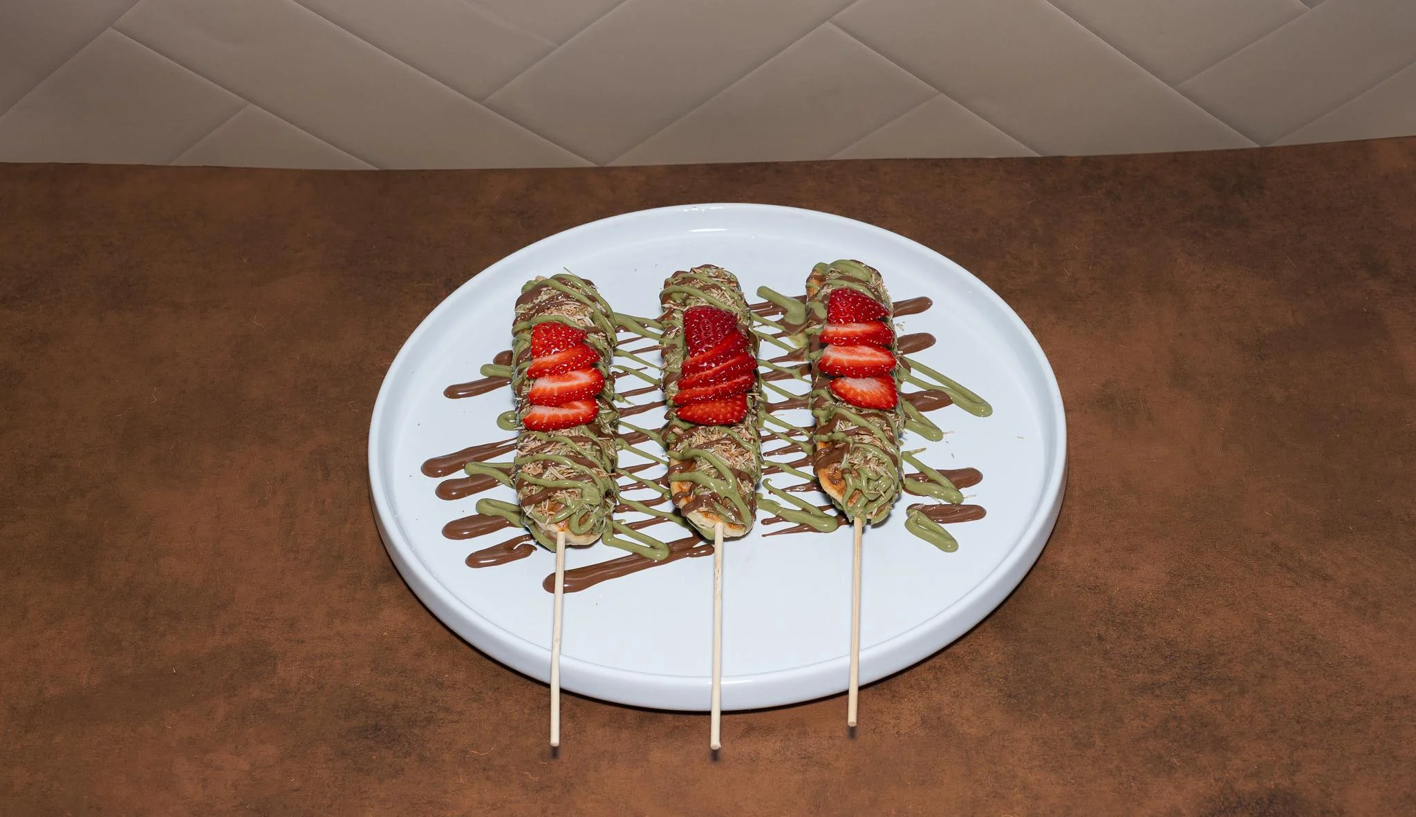 Three skewered treats on a white plate, decorated with green and brown sauces, topped with fresh strawberries.