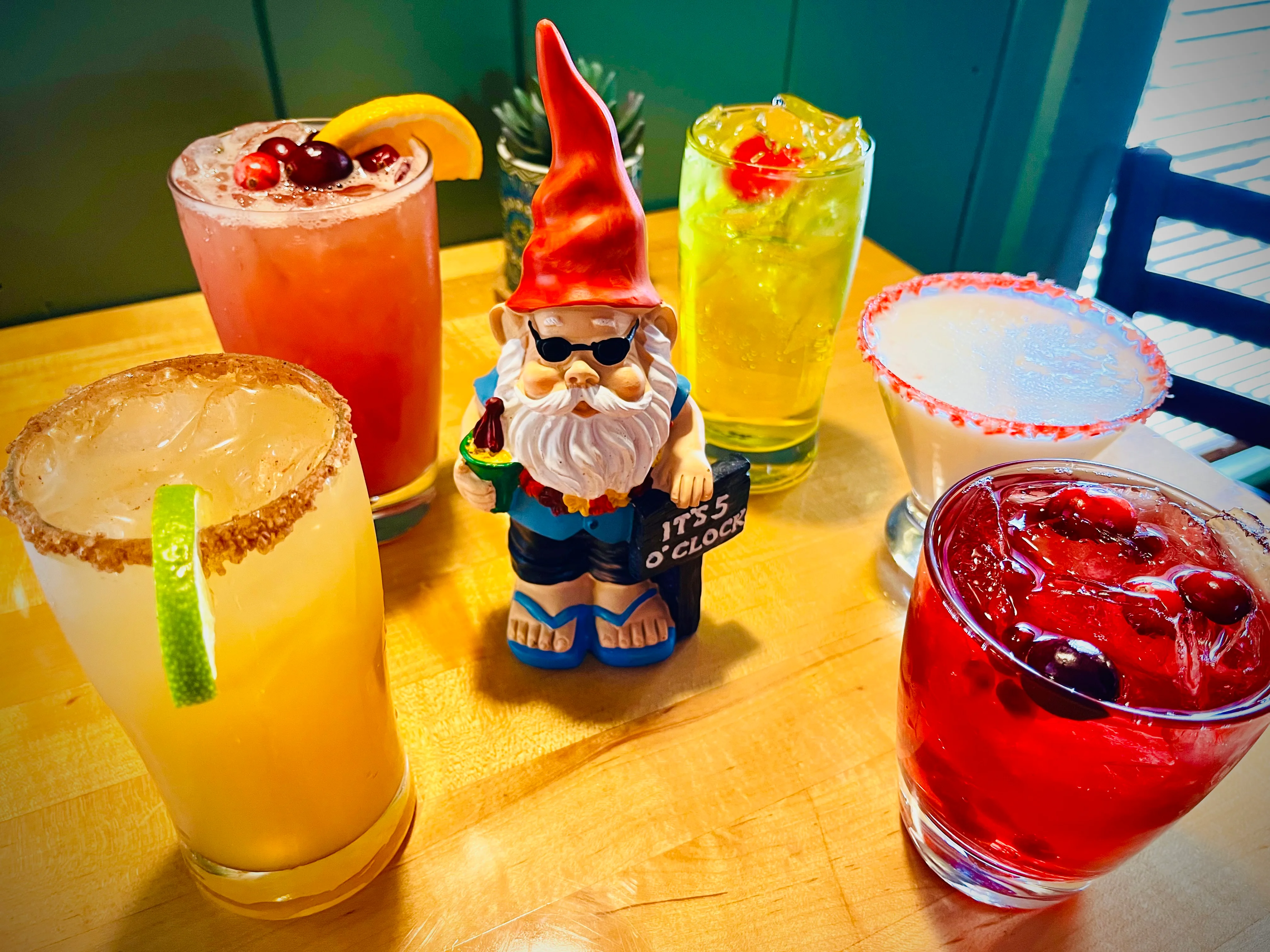 A colorful array of cocktails arranged around a cheerful gnome figurine, emphasizing fun and festive drinks.