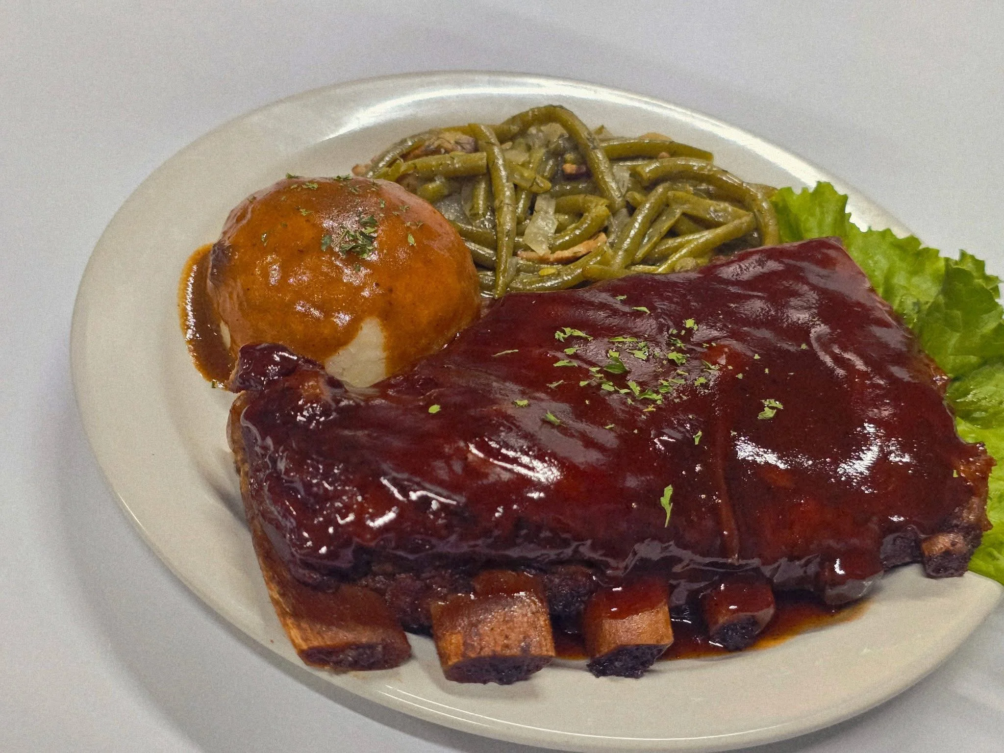 A hearty plate featuring barbecue ribs glazed with sauce, served alongside mashed potatoes and green beans, garnished with lettuce.