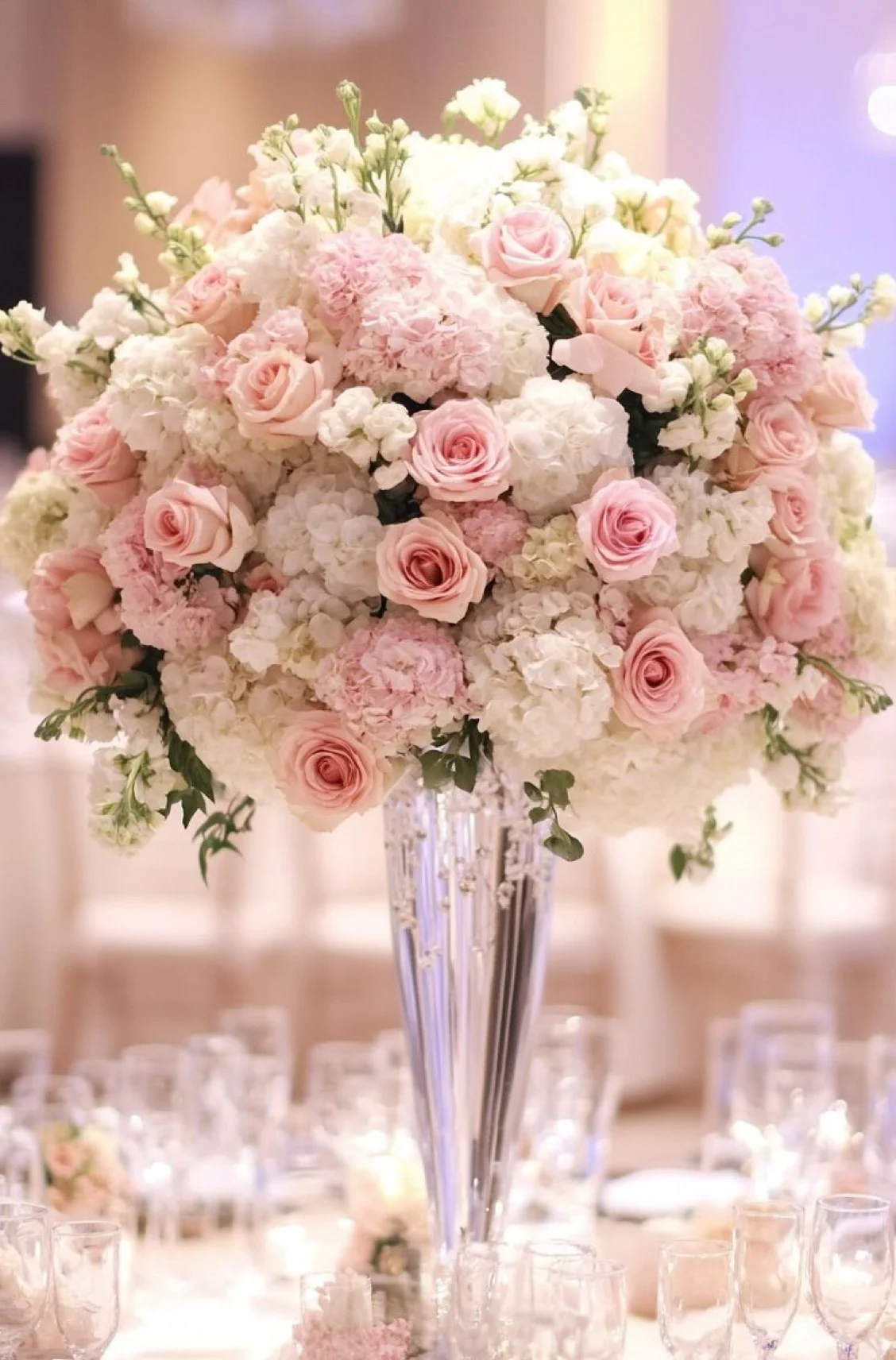 An elegant floral arrangement featuring pink roses and white hydrangeas in a tall, crystal vase, surrounded by dining settings.