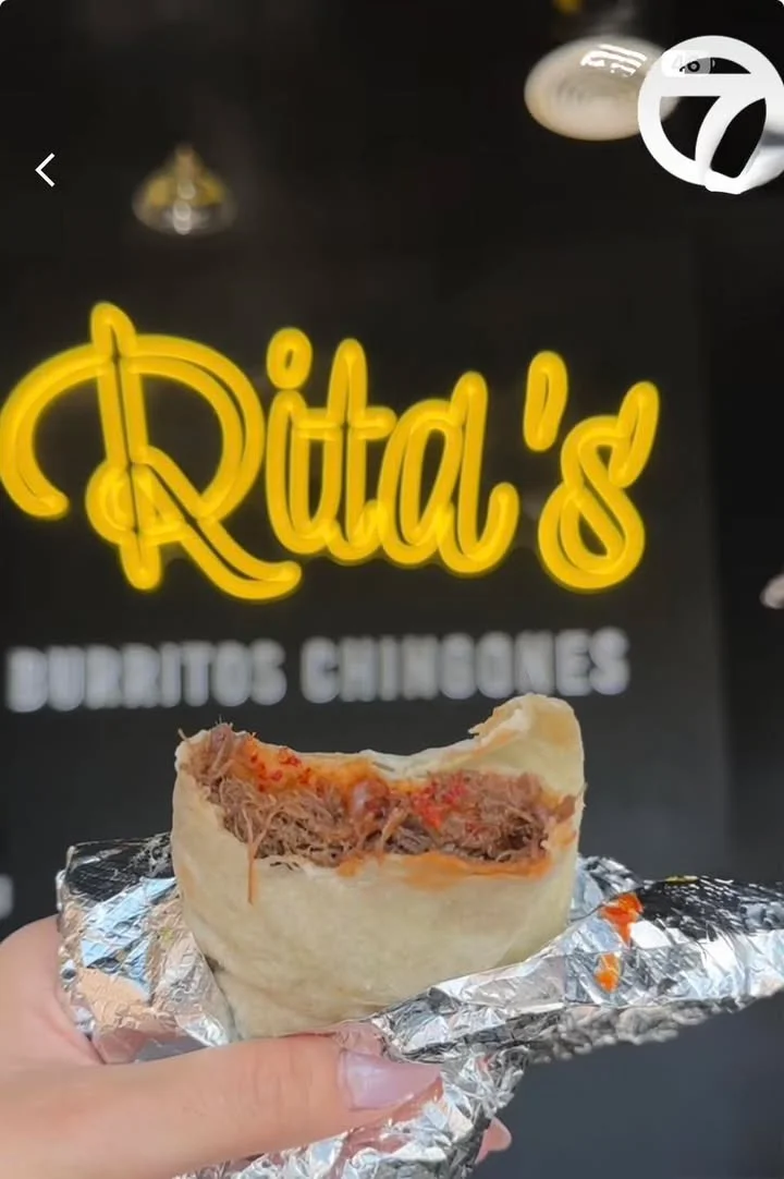 A person holding a burrito showing its filling, with a brightly lit sign in the background.