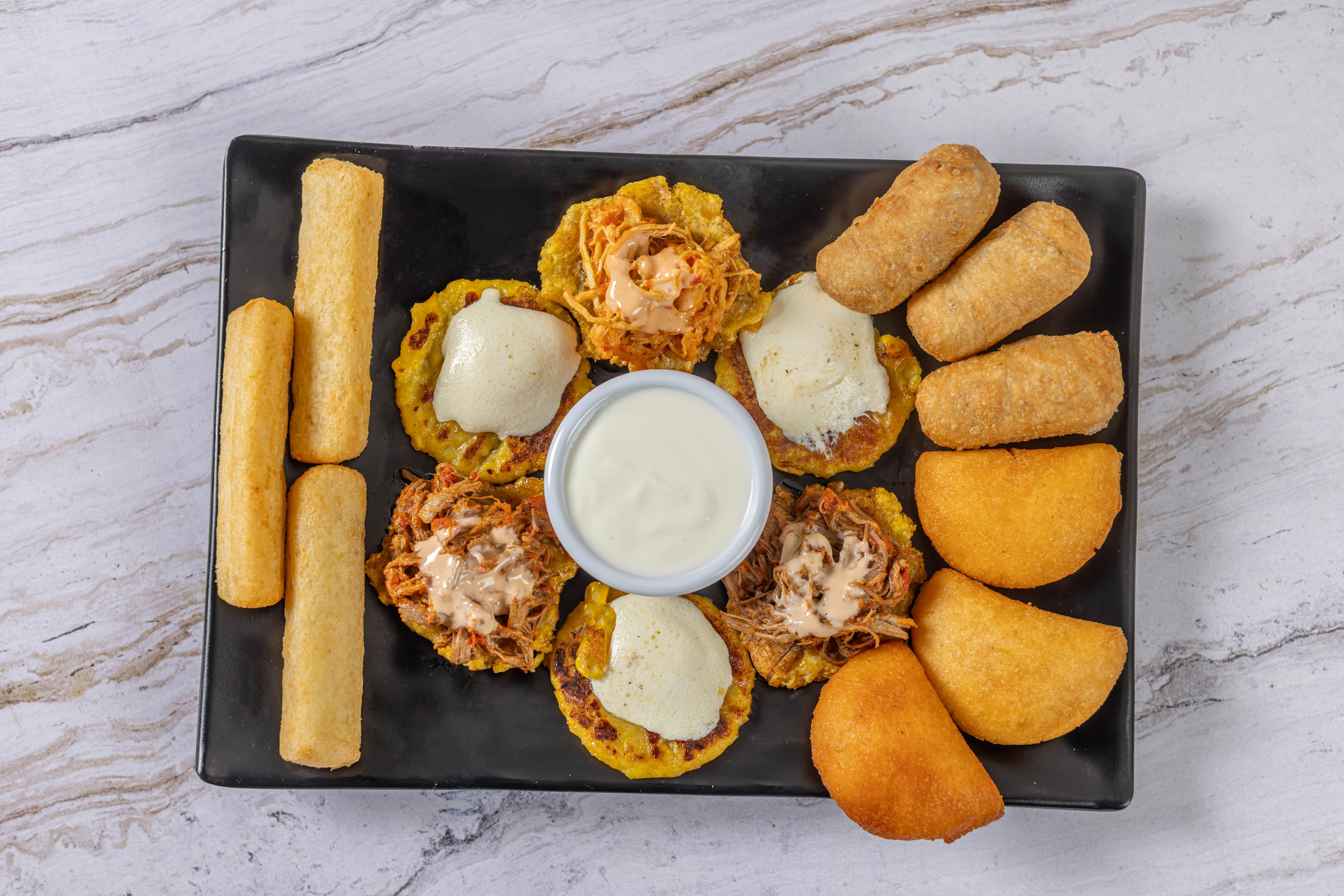 An appetizer platter of classic Latin starters, featuring tequeños (cheese sticks), mini empanadas, and tostones rellenos with a creamy dipping sauce.