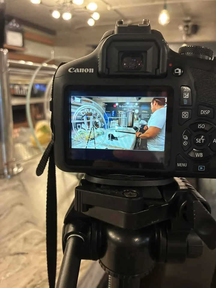 A camera on a tripod displays a photo of a countertop with a distillation apparatus. The scene features a person engaged with the setup in a well-lit indoor space.