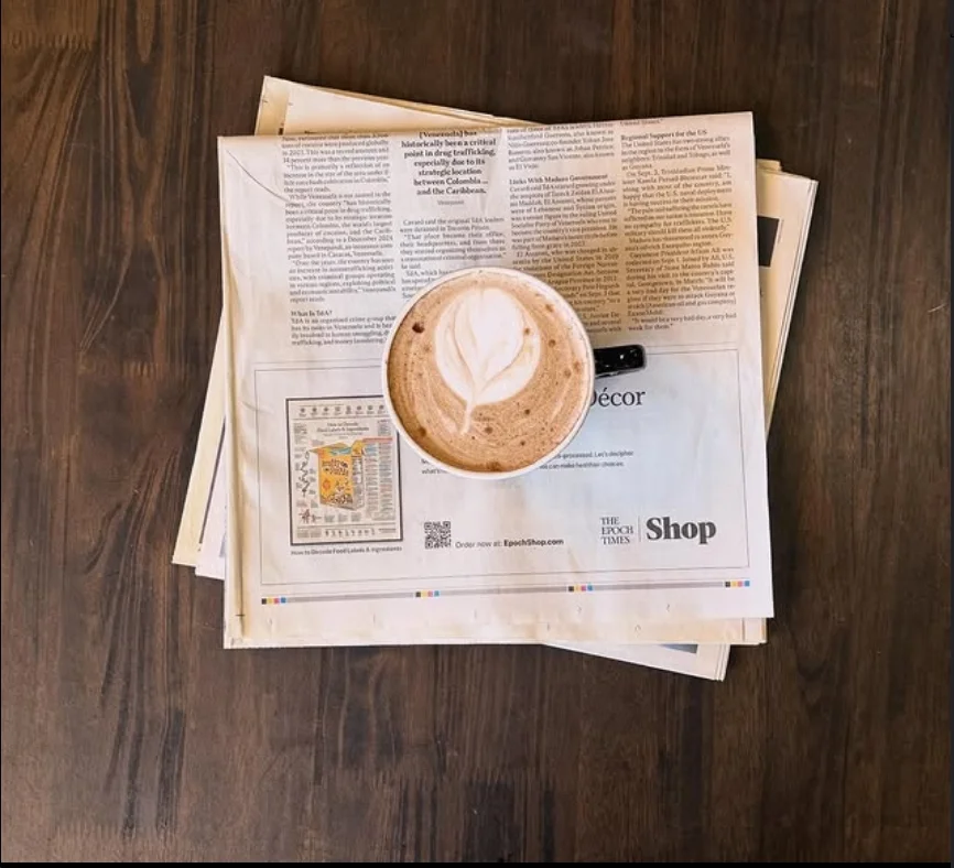A cup of coffee with latte art on top, placed on a stack of newspapers on a wooden table.