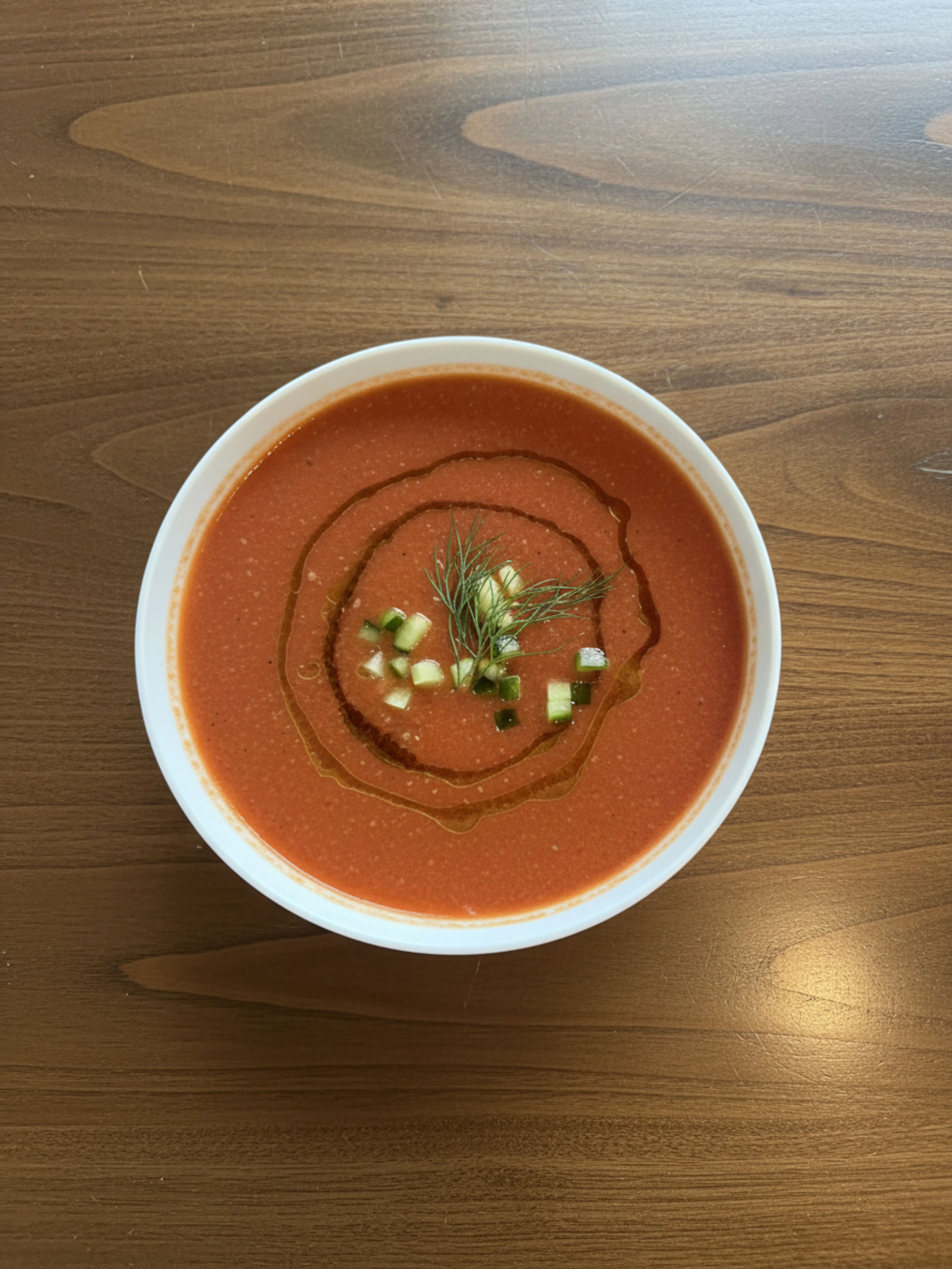 A bowl of vibrant red tomato soup drizzled with olive oil, garnished with diced cucumbers and fresh herbs.