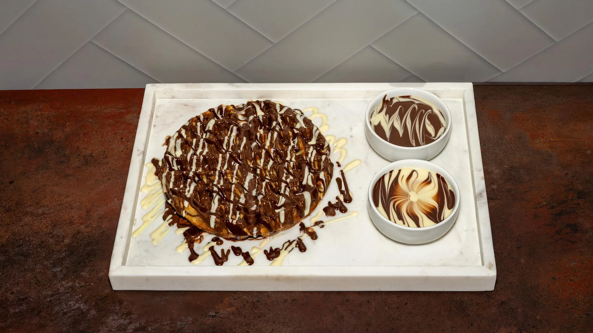A round chocolate cake drizzled with white icing, accompanied by two bowls of creamy chocolate sauce, served on a marble tray.