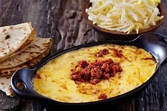 A serving of melted cheese topped with chorizo, accompanied by warm tortillas and shredded cheese on the side.
