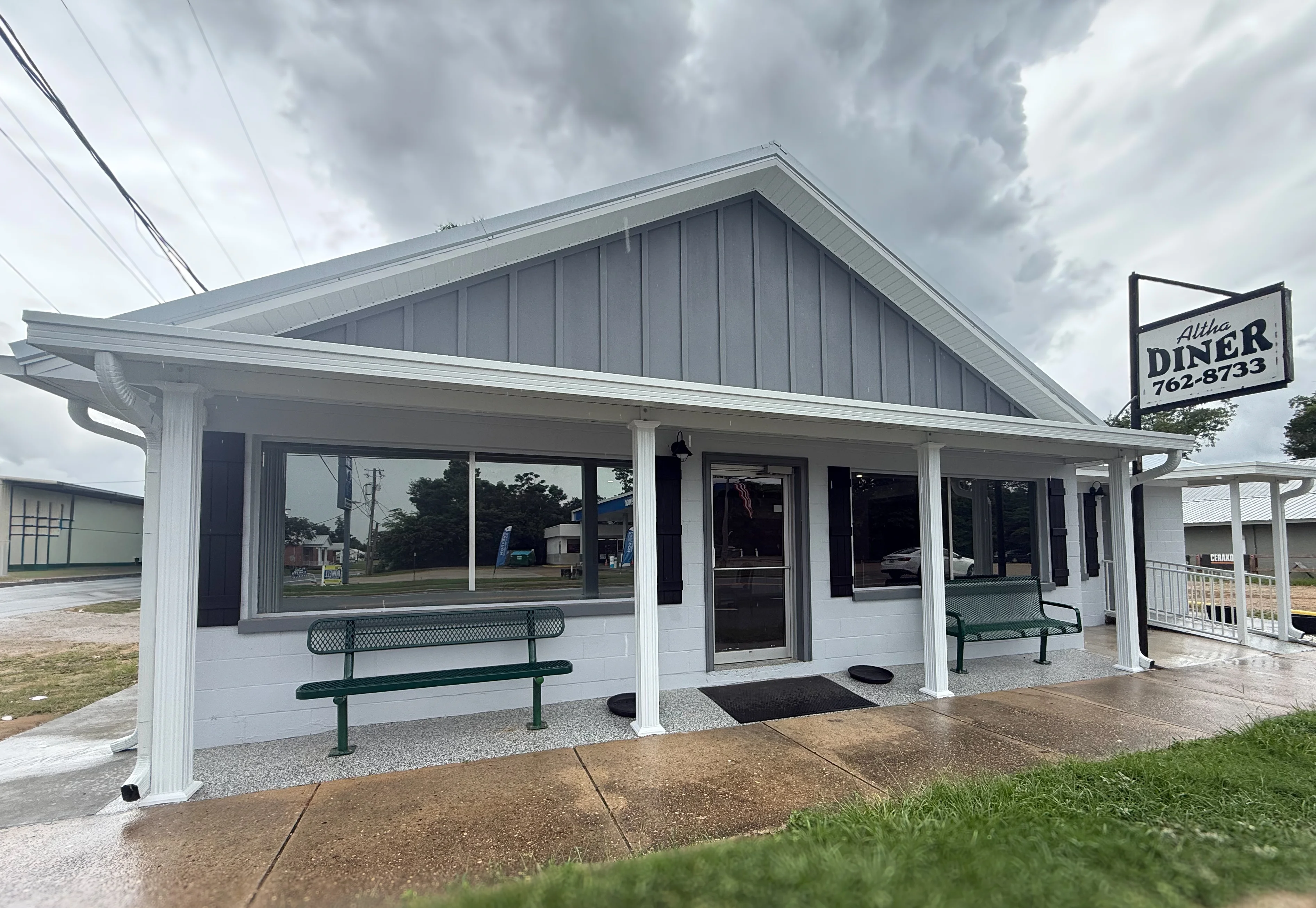 A diner exterior featuring a grey and white building with a signage that includes the name 'Altha Diner'. The front has a covered area with a green bench and large windows.