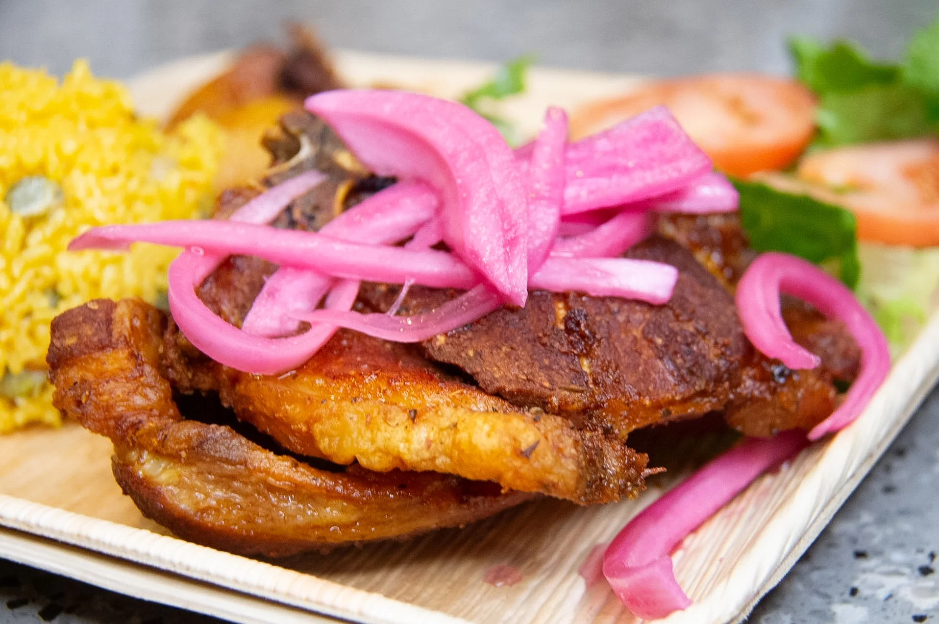A plate featuring a crispy chicken piece topped with pink onions, served alongside yellow rice and fresh tomato slices.
